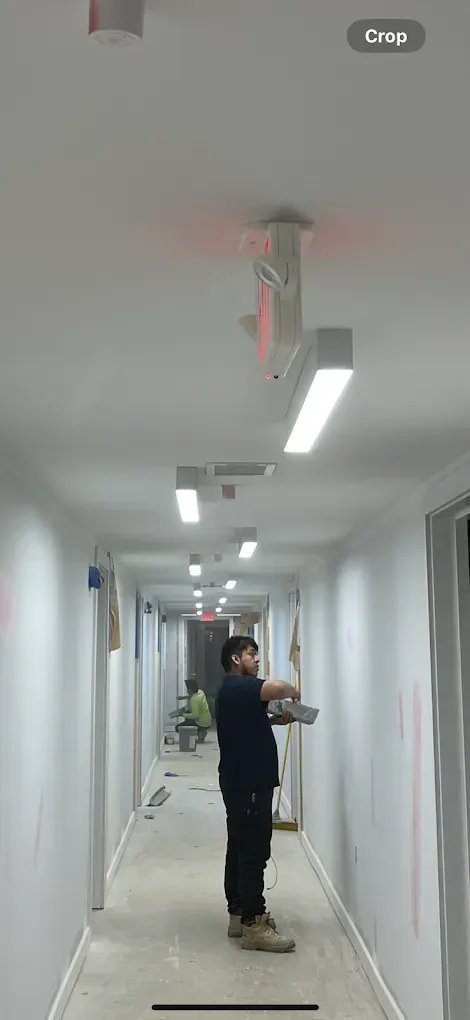 A person works in a long, white hallway, holding a metal tray. Fluorescent lights and a red-lit object are on the ceiling.