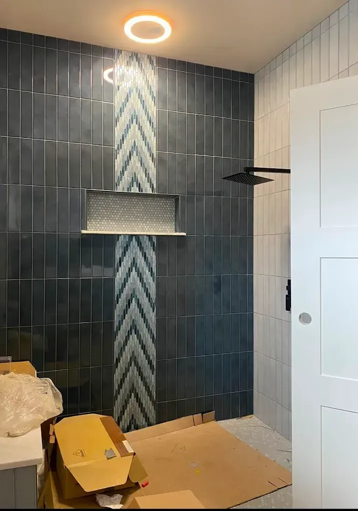 Bathroom shower with blue tiles, patterned accent, shelf, black showerhead, and white door.