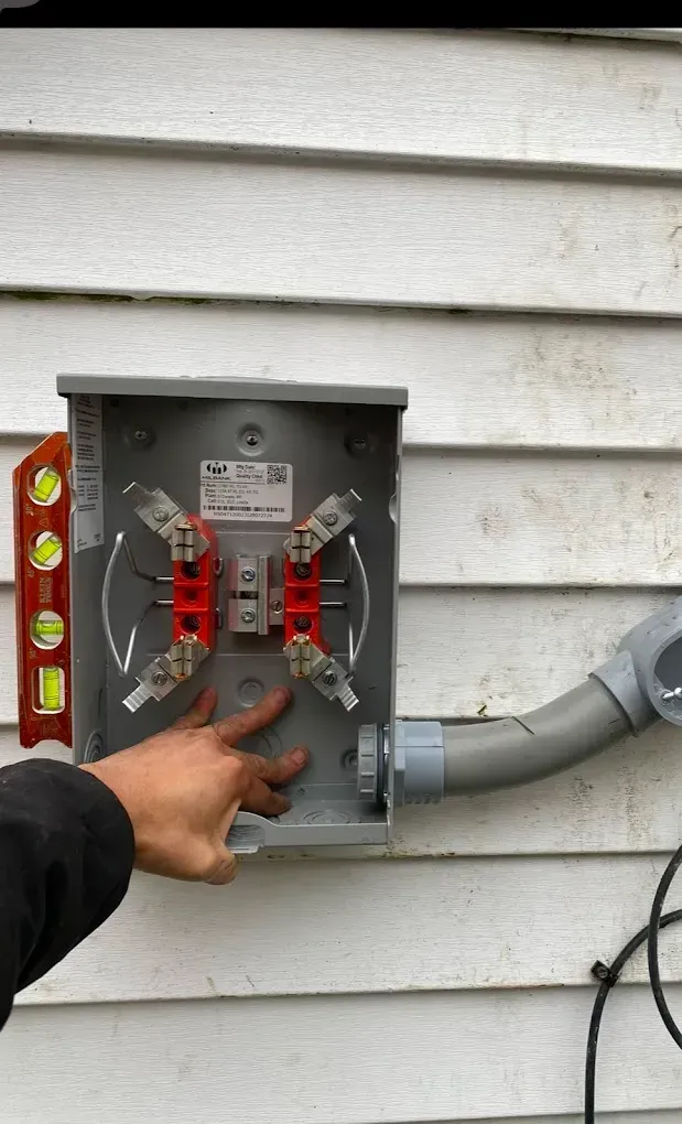 A person points to an open electrical box attached to a house with conduit. A level is nearby.