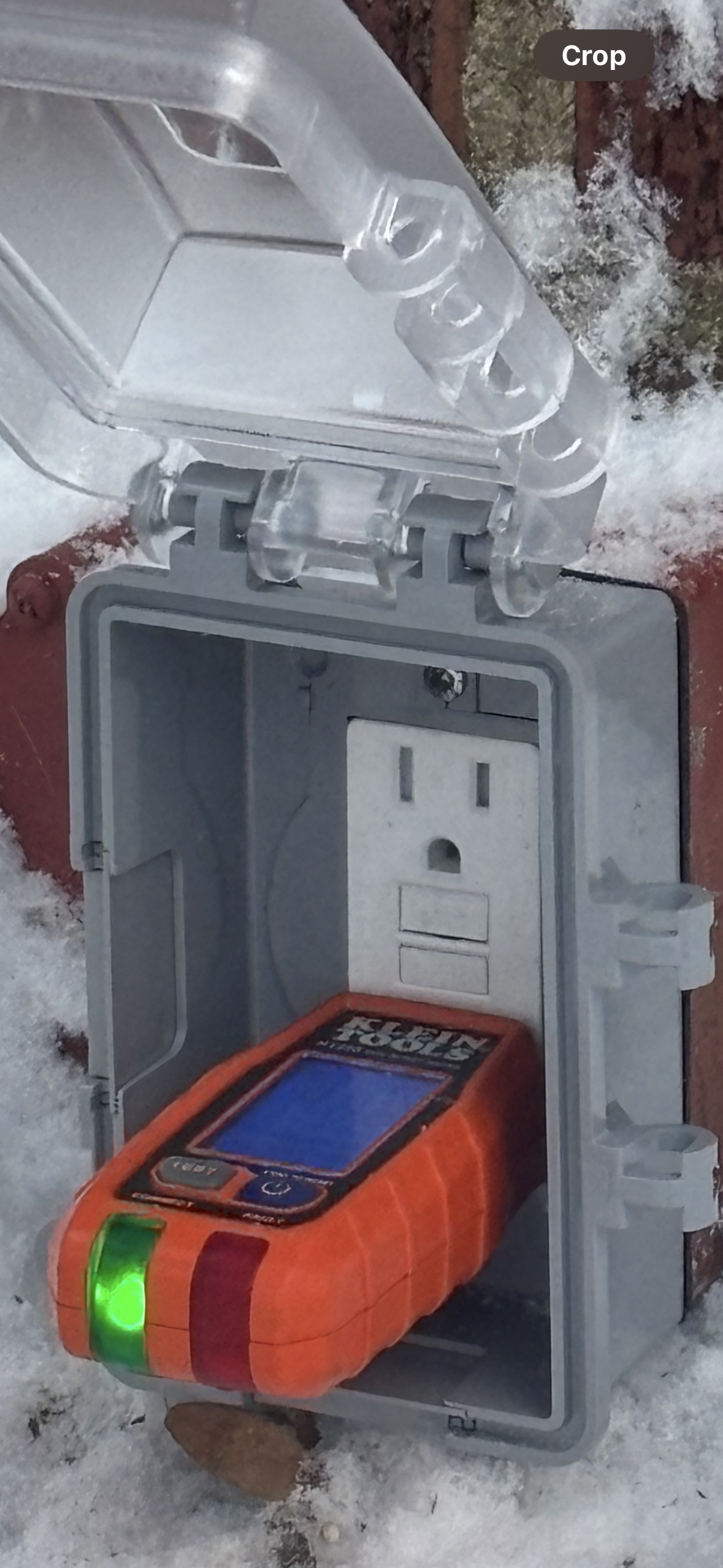 An orange electrical tester plugged into a power outlet within a gray weatherproof box. The tester's light is green.