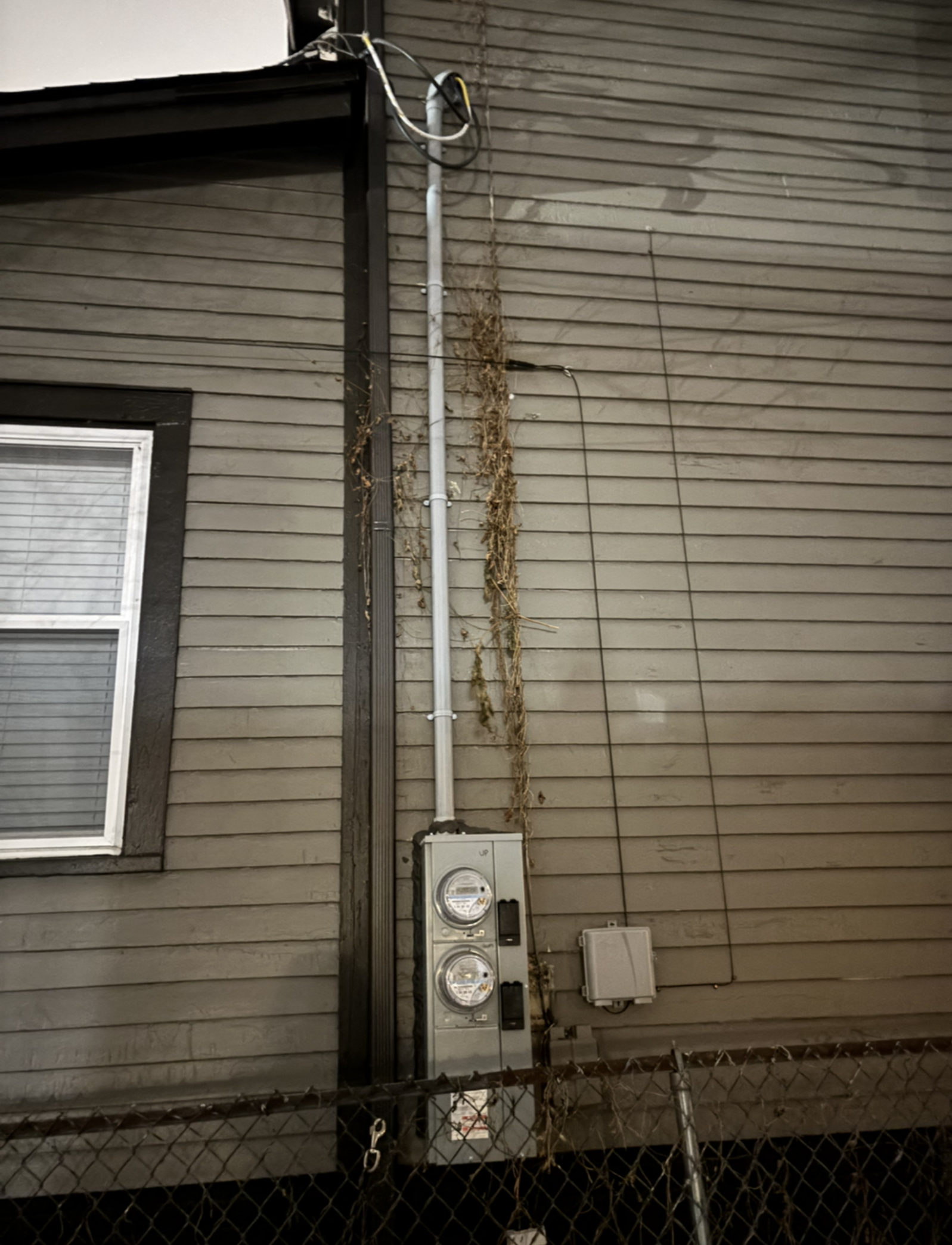 Electrical equipment attached to a building, including a meter box and conduit, with a chain-link fence below.