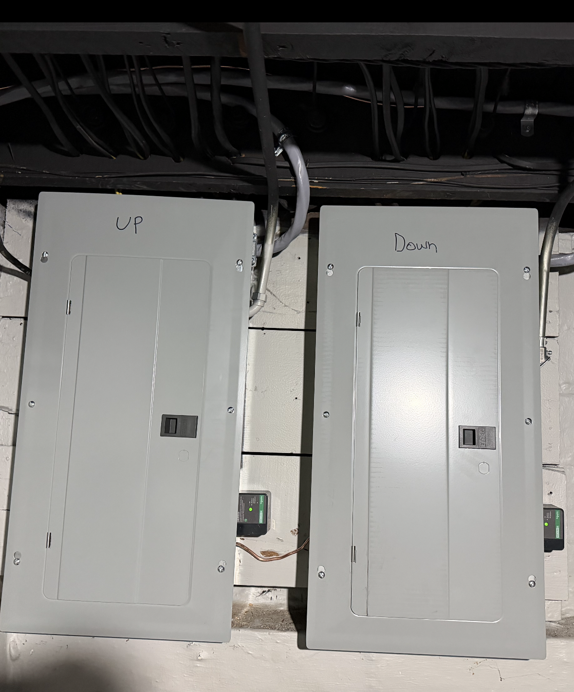 Two closed gray electrical panels mounted on a brick wall. Electrical wiring overhead.