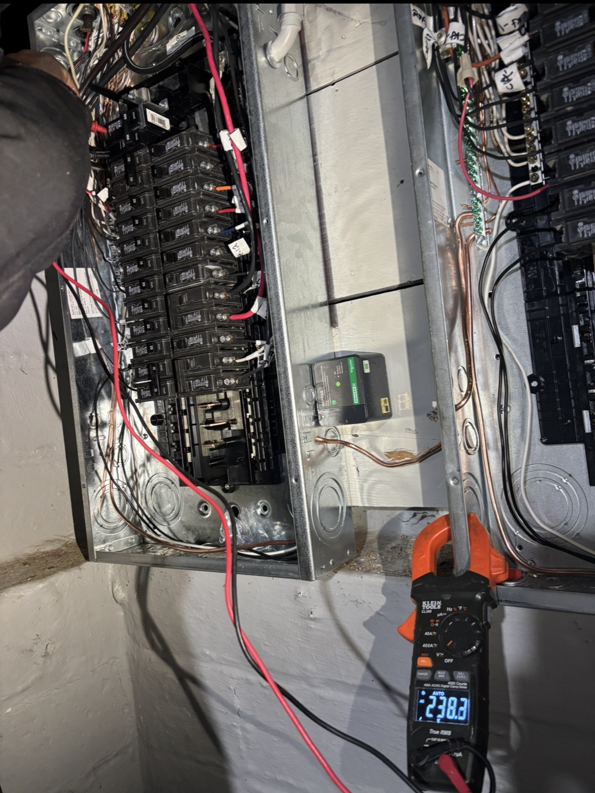An electrician using a clamp meter on electrical panel. Red and black wires are visible. Meter reads 27.52.