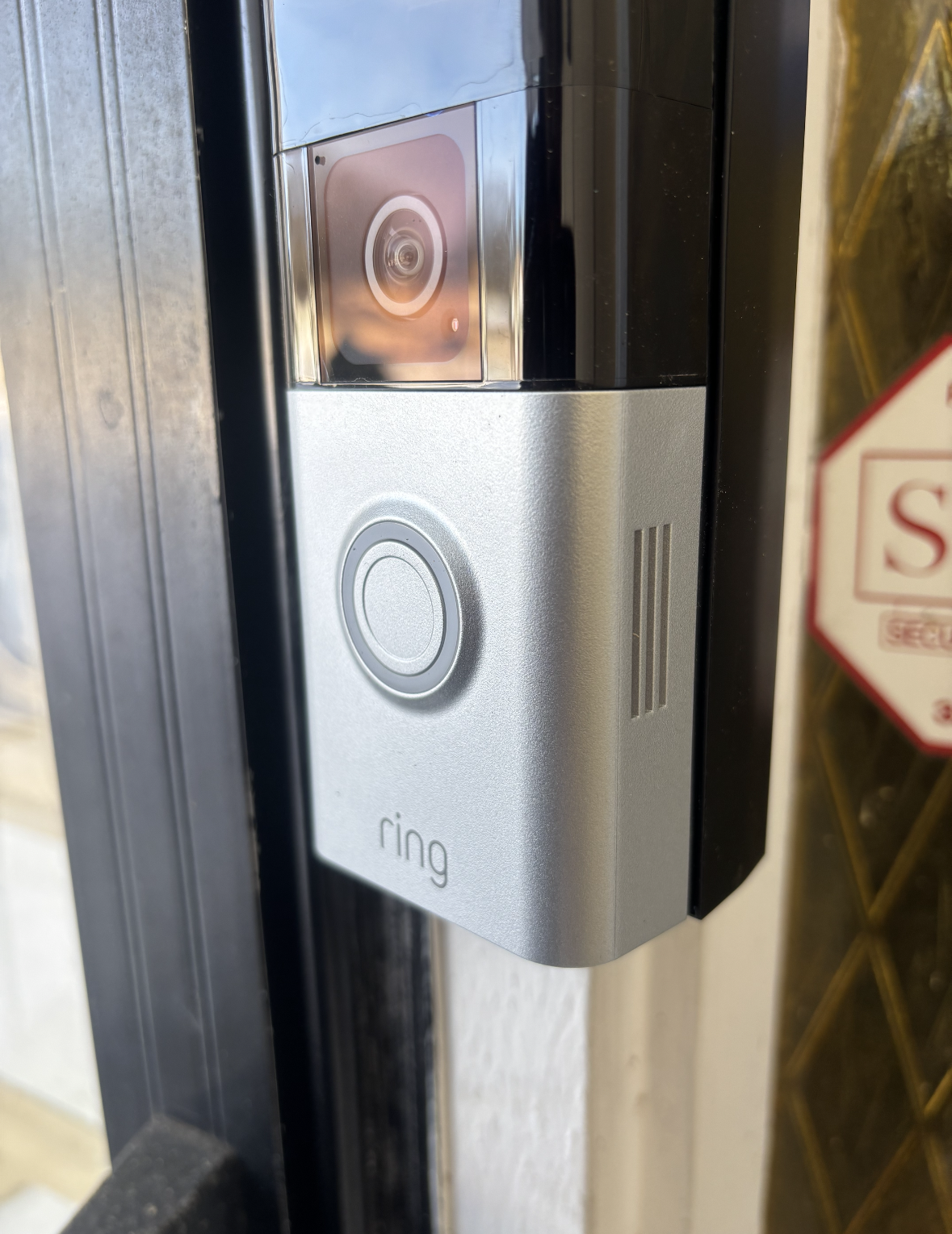 Silver Ring video doorbell mounted on a black door frame. The doorbell has a button and camera.