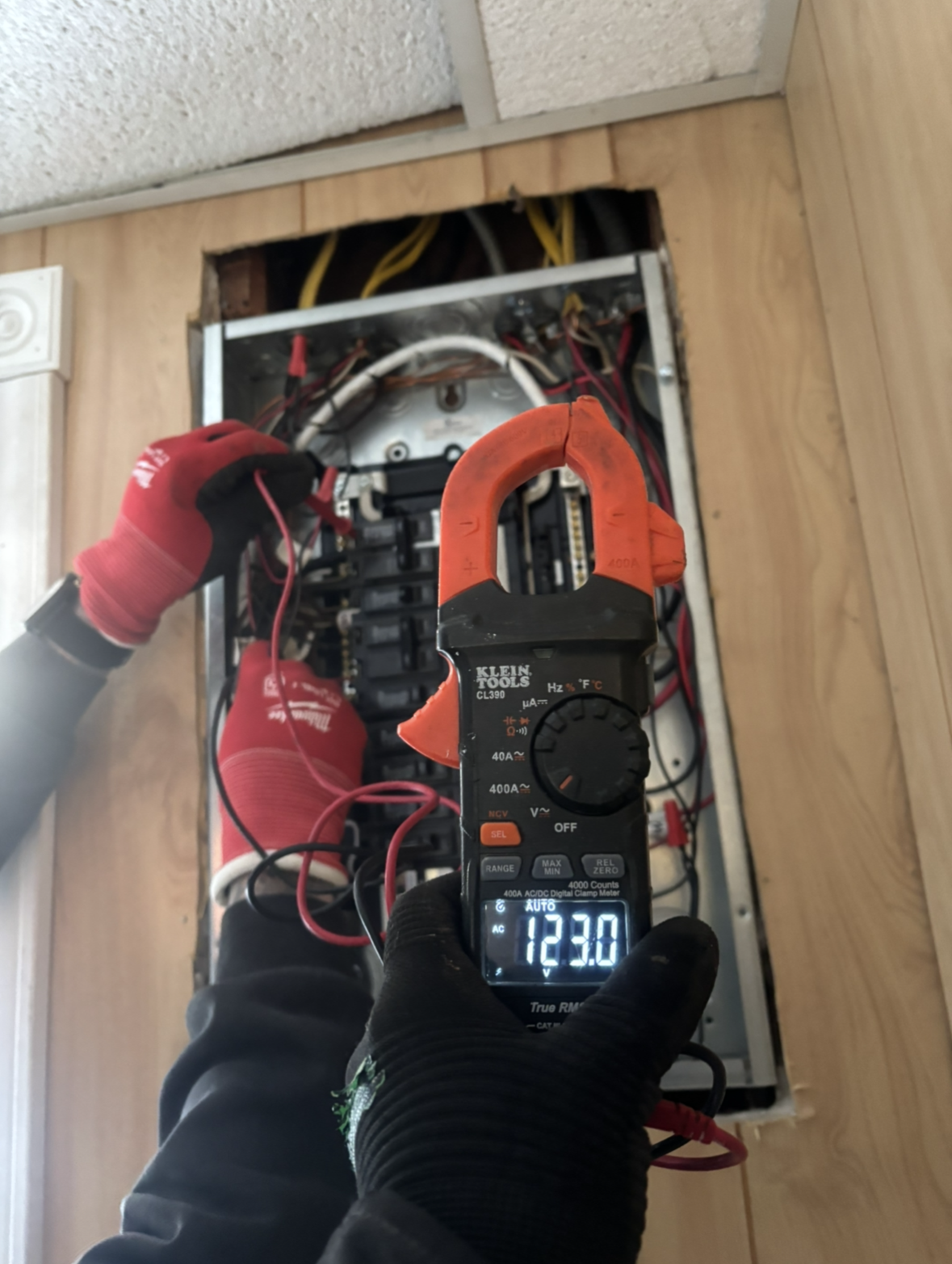 An electrician uses a clamp meter to measure electrical current in a breaker panel. The meter reads 12.30.