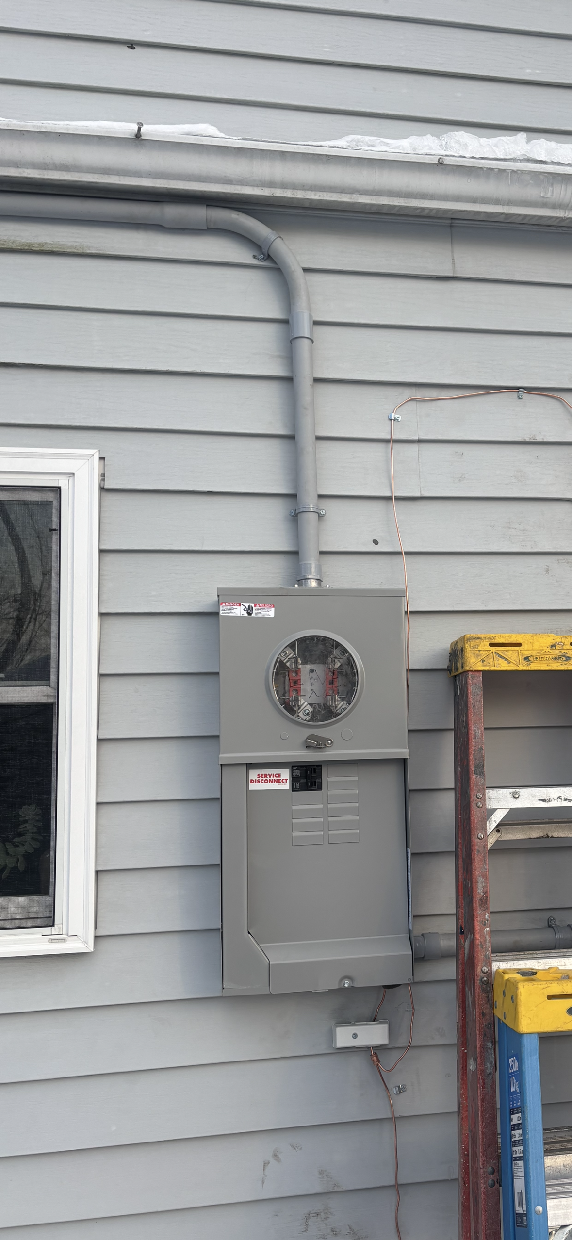 Gray electrical box mounted on a gray siding wall, with a window on the left and a ladder on the right.