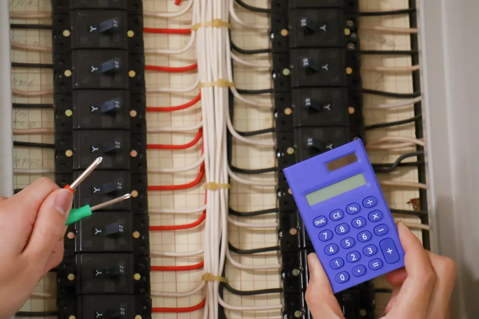 Hands holding wires and a calculator near an open electrical panel.