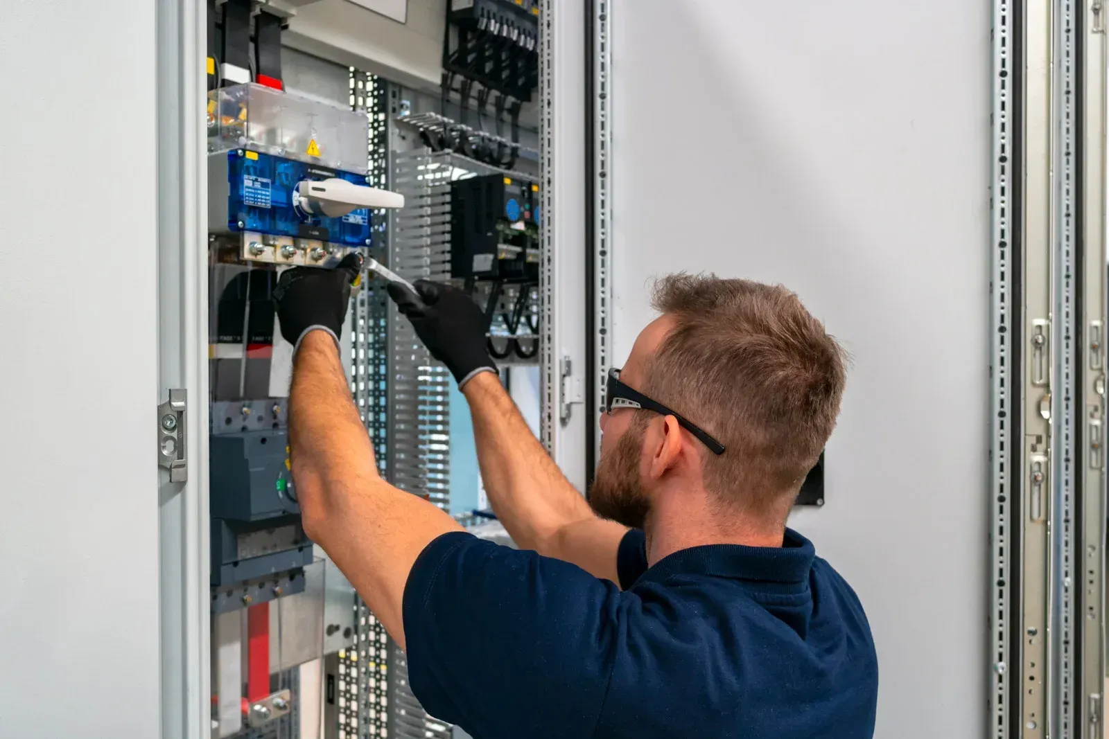 Electrician in black gloves and safety glasses works inside an electrical panel.