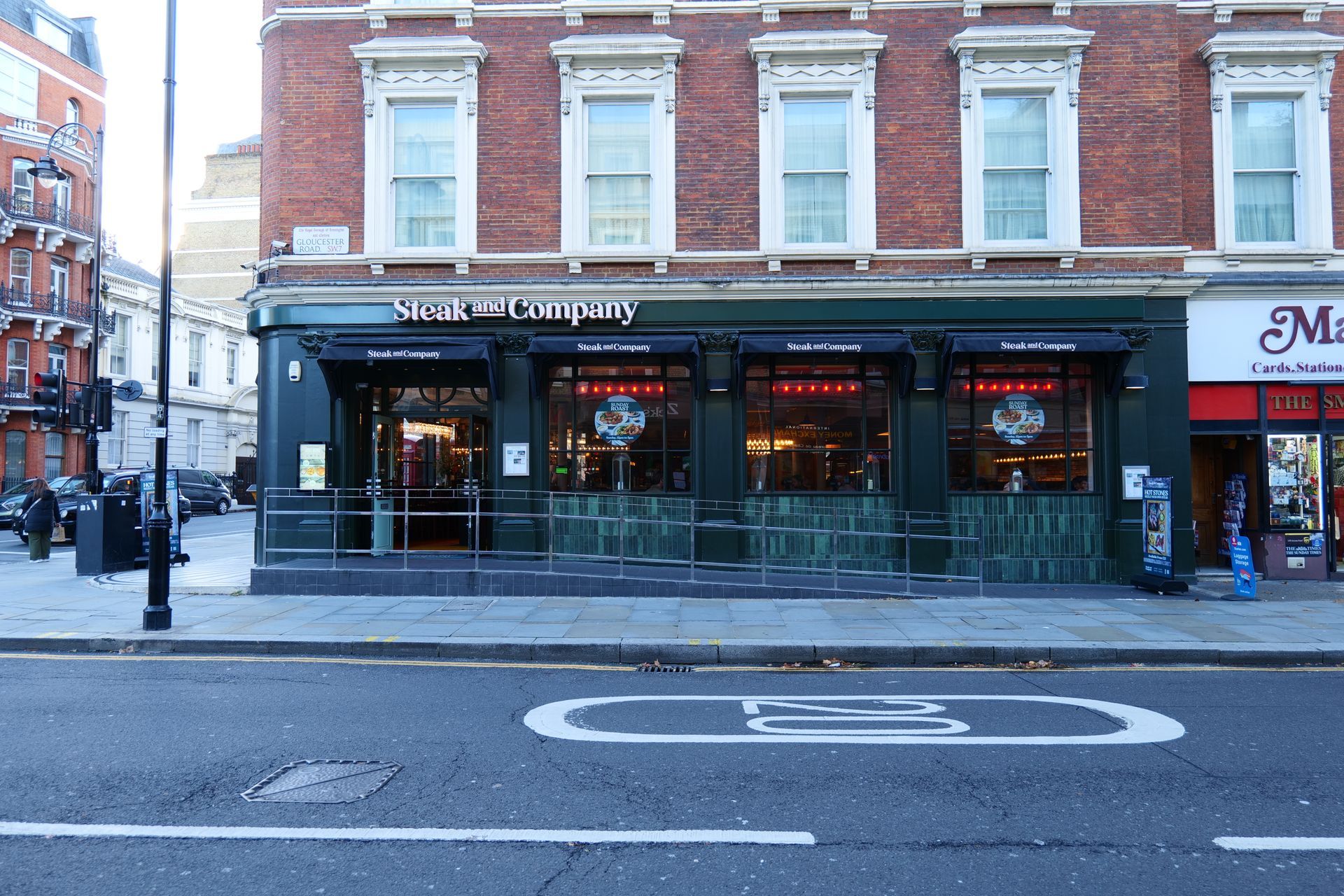 Steak and Company restaurant exterior. Green facade, large windows, outdoor seating, and a busy street scene.