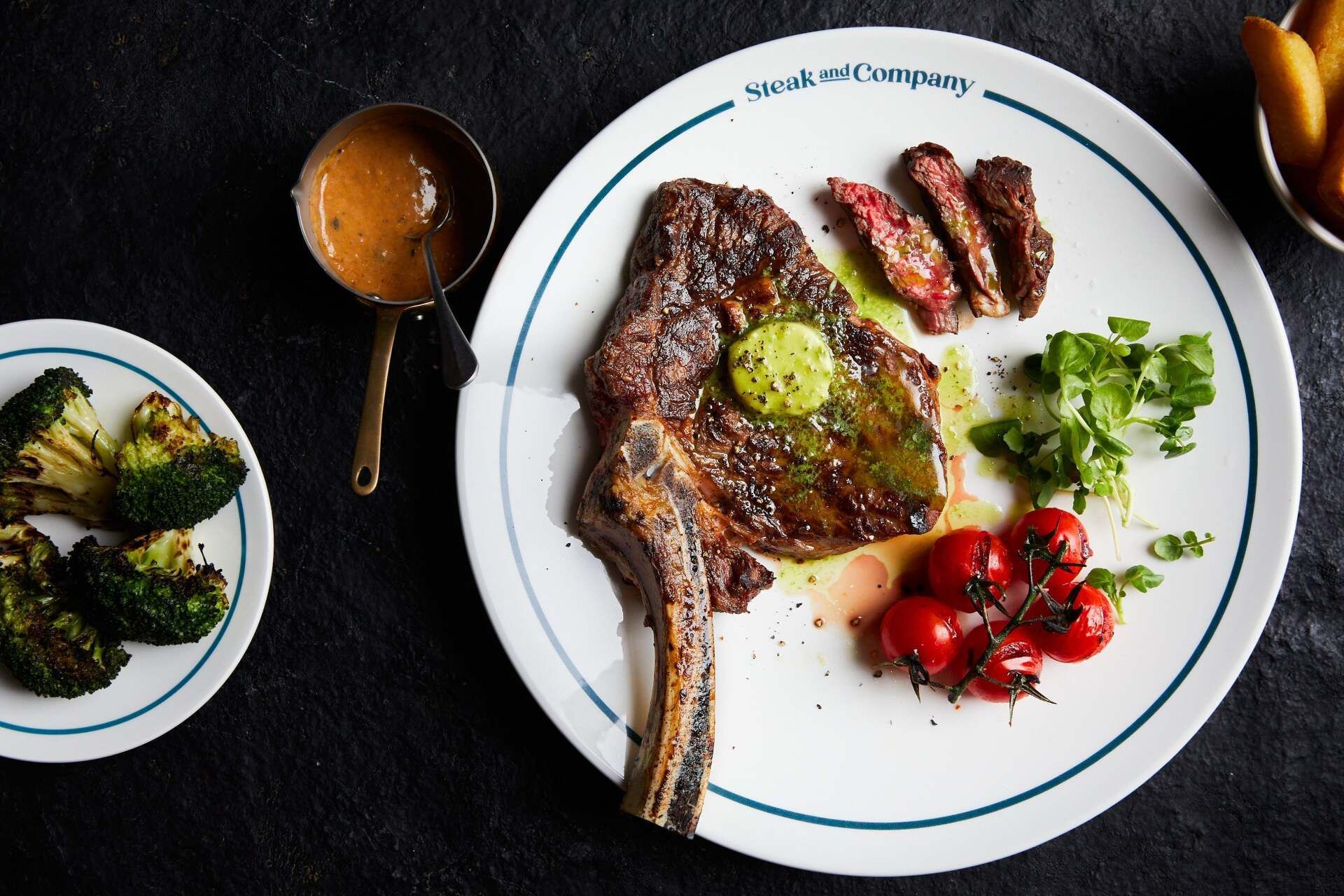 Plate of steak with side dishes: broccoli, tomatoes, sauce, and french fries.