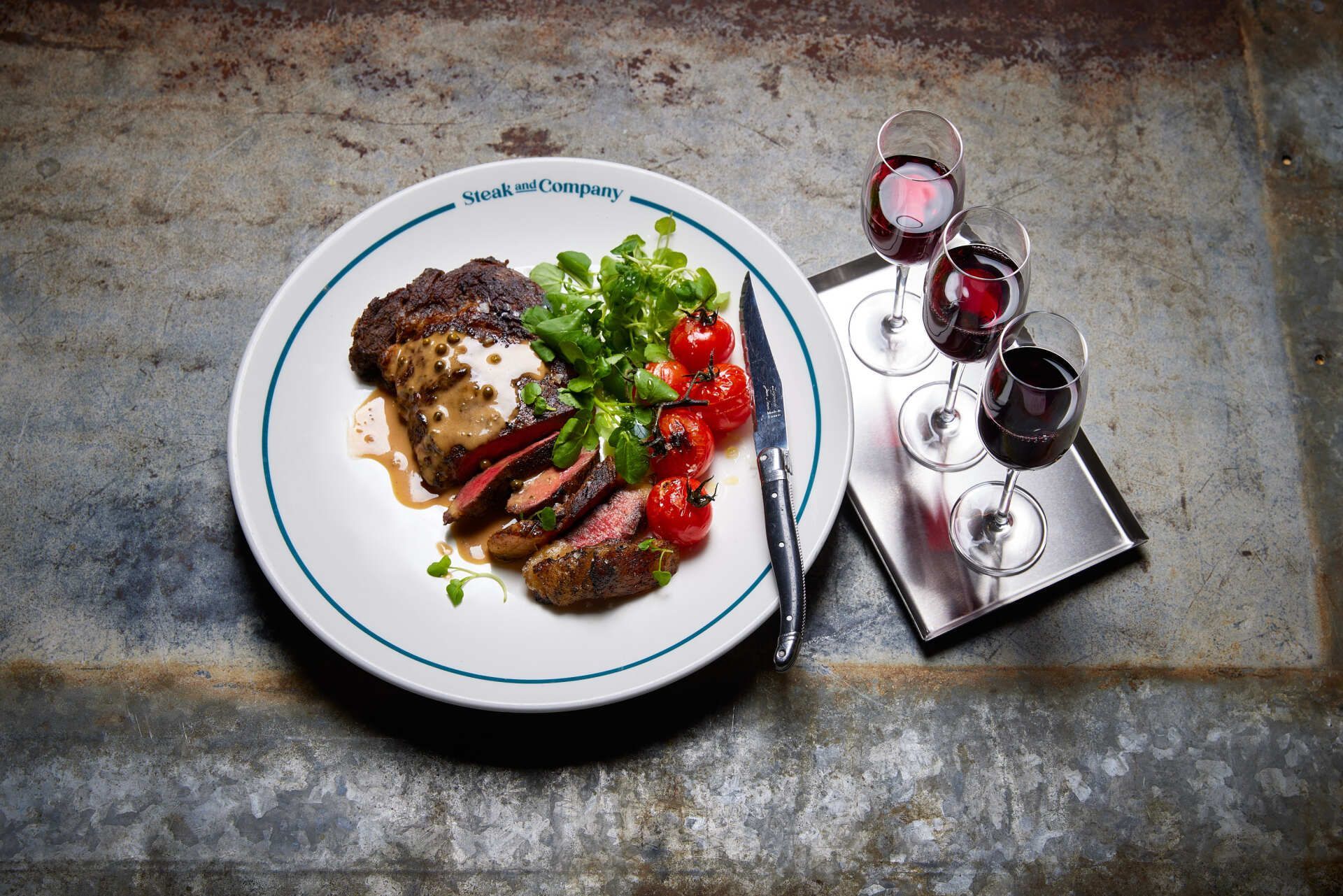 Steak with tomatoes and greens on a plate, with red wine glasses on a tray.