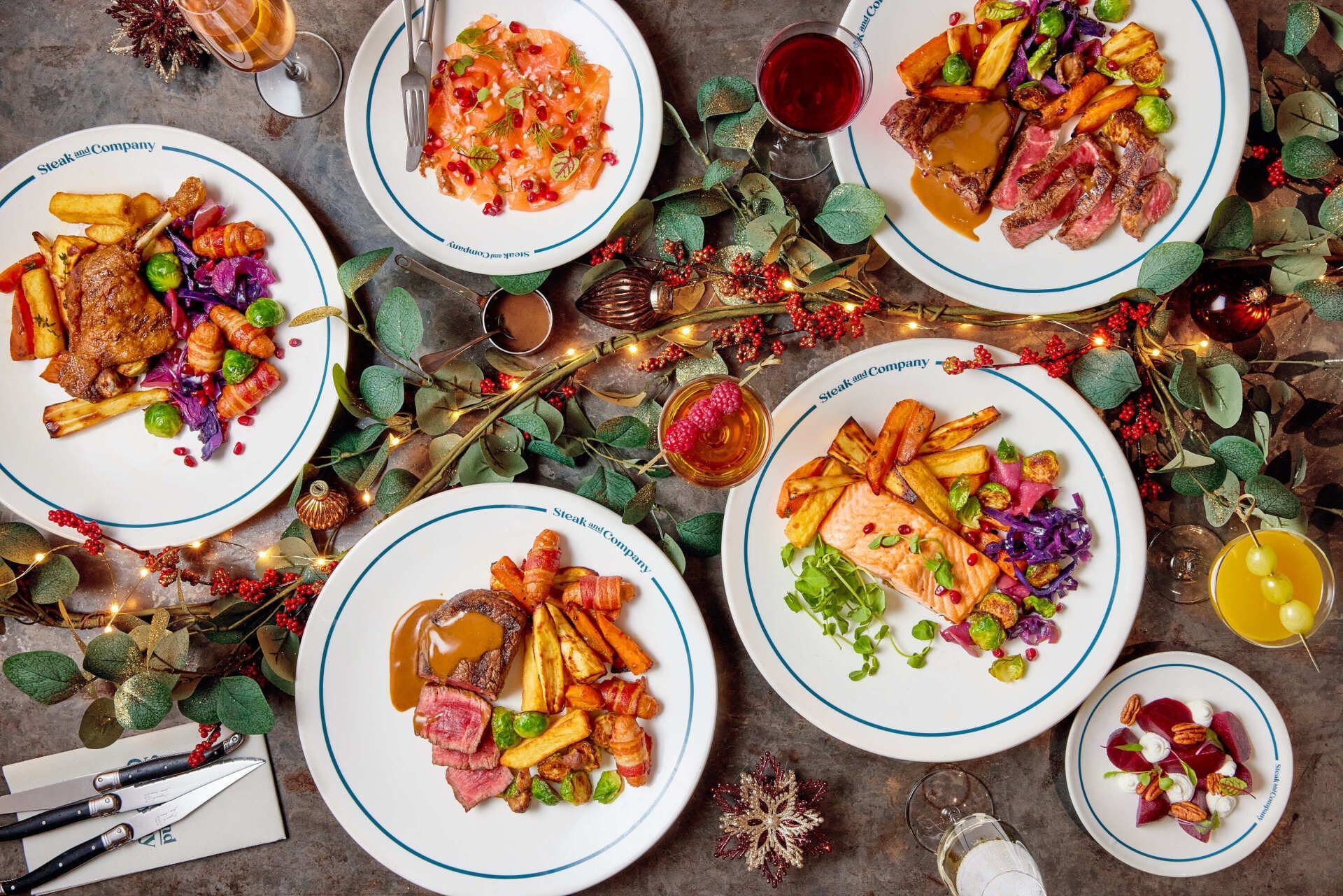 Plates of food on a table with festive decorations including greens, lights, and drinks.