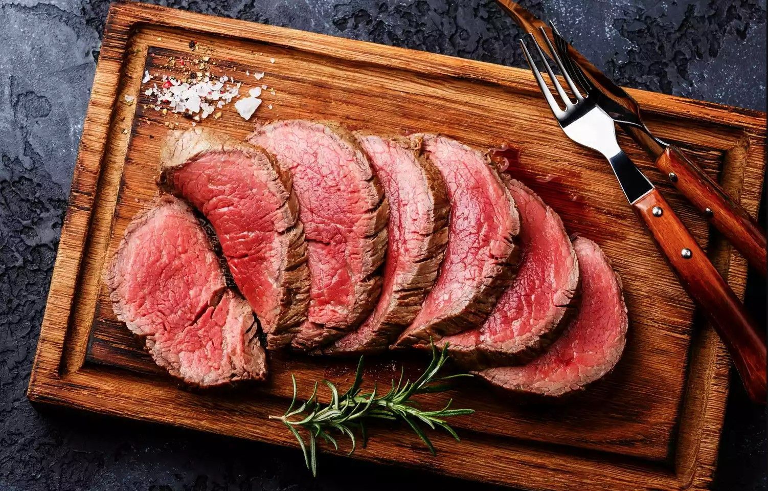 Sliced, medium-rare roast beef on a wooden board with salt, rosemary, and utensils.