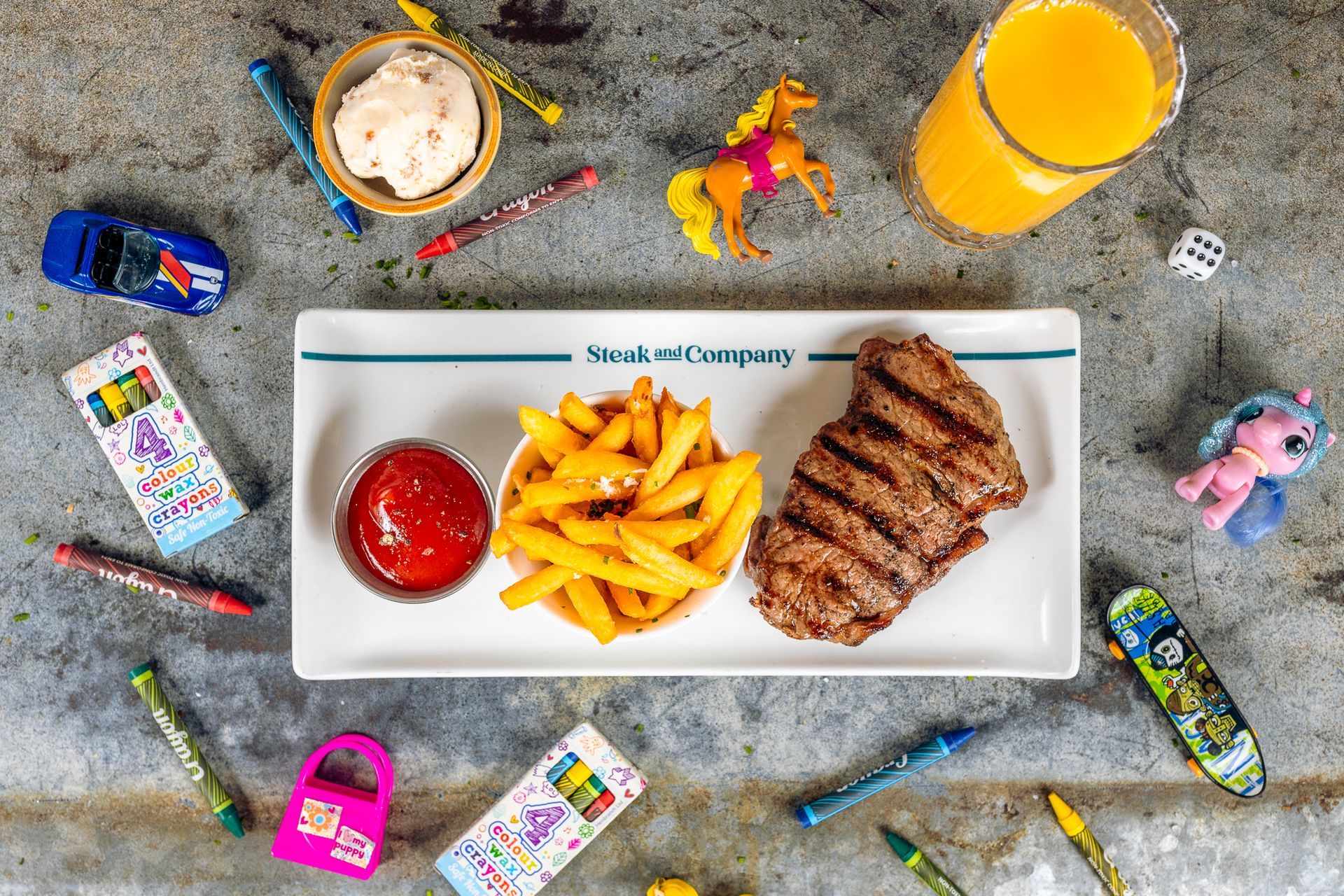 Steak, fries, ketchup, and orange juice on a tray with toys and crayons scattered around.
