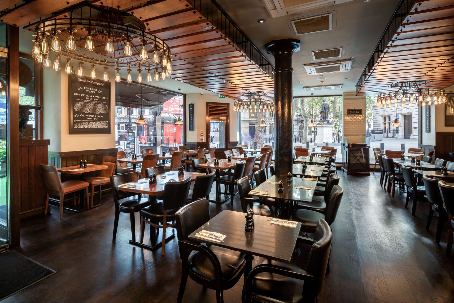 Restaurant interior with tables, chairs, and ornate light fixtures; wooden floors and ceiling, windows view street.