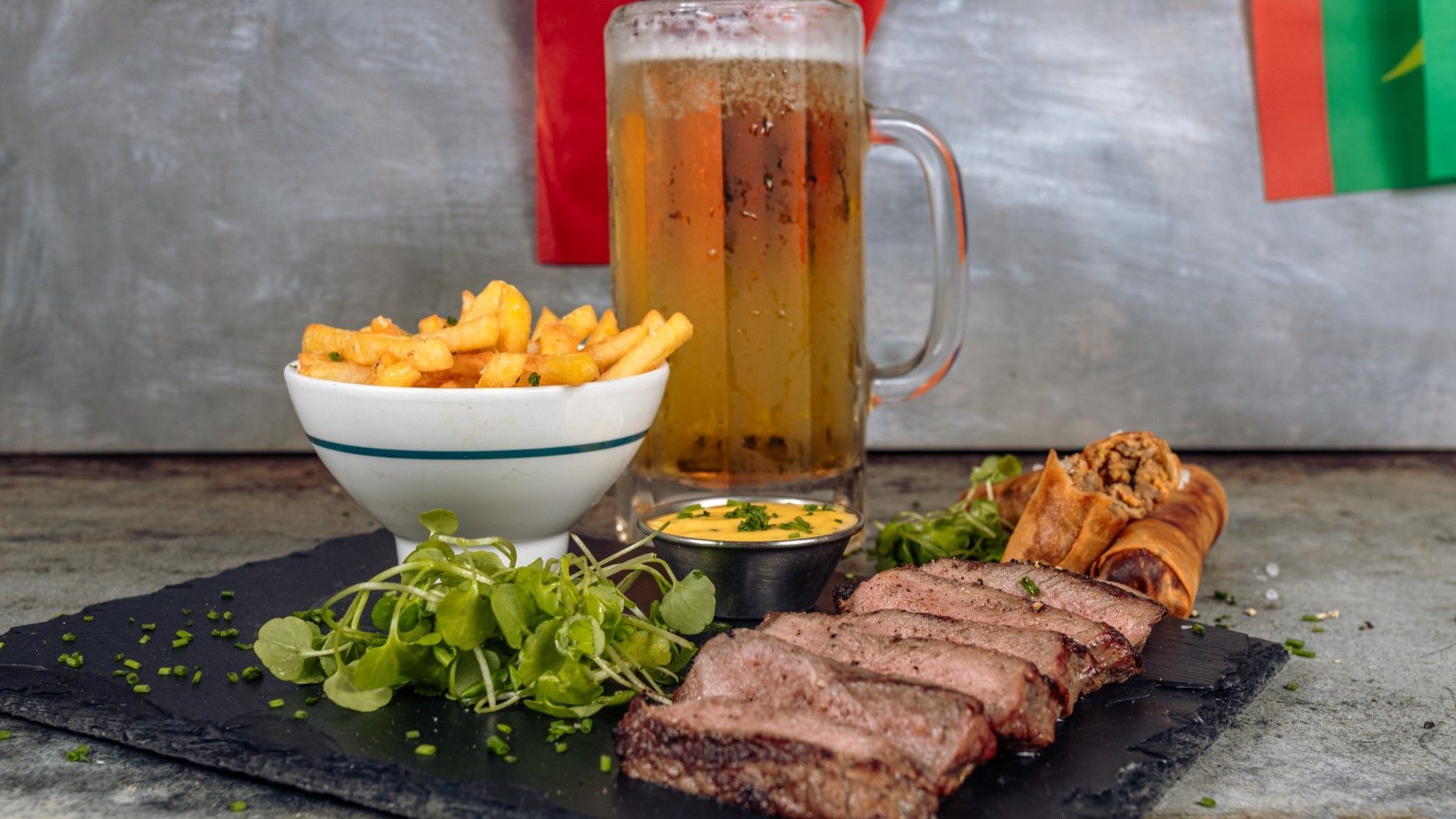 Steak, fries, beer in a mug, and dipping sauce arranged on a slate board.