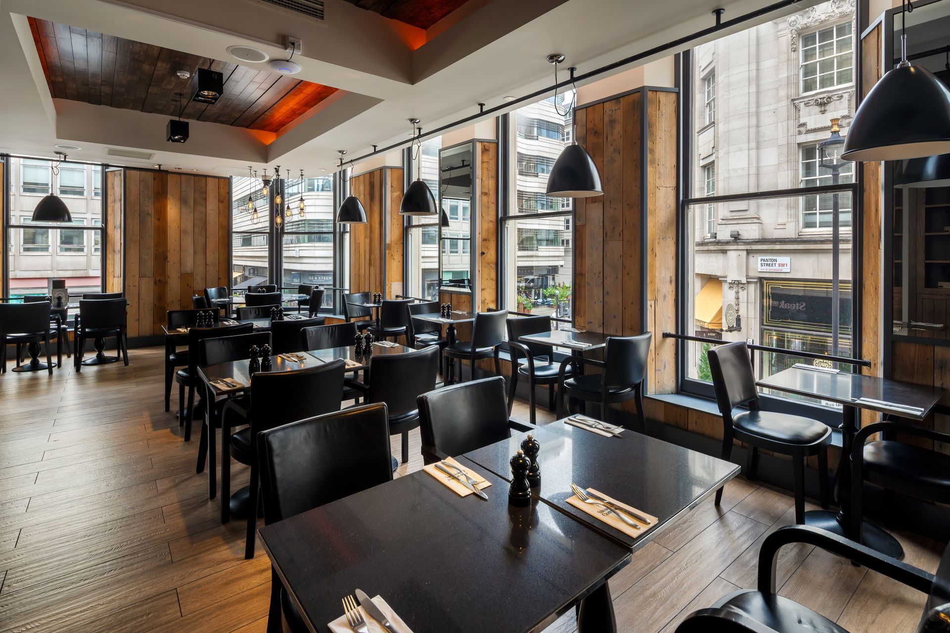 Restaurant interior with dark tables and chairs, large windows, and wood paneling.