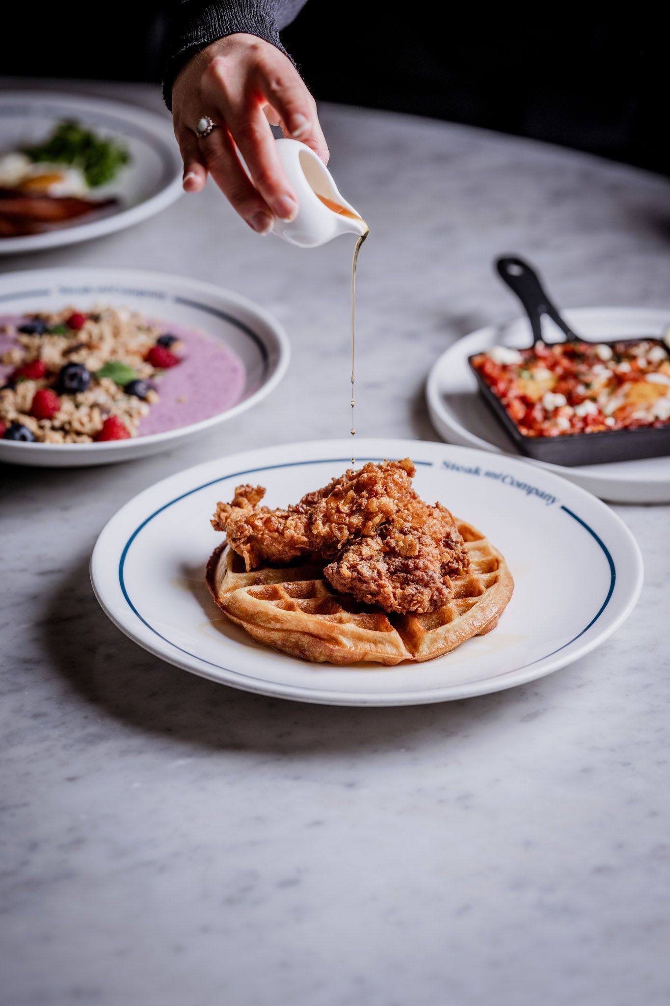 Chicken and waffle dish with syrup being poured, on a marble table with other breakfast plates.