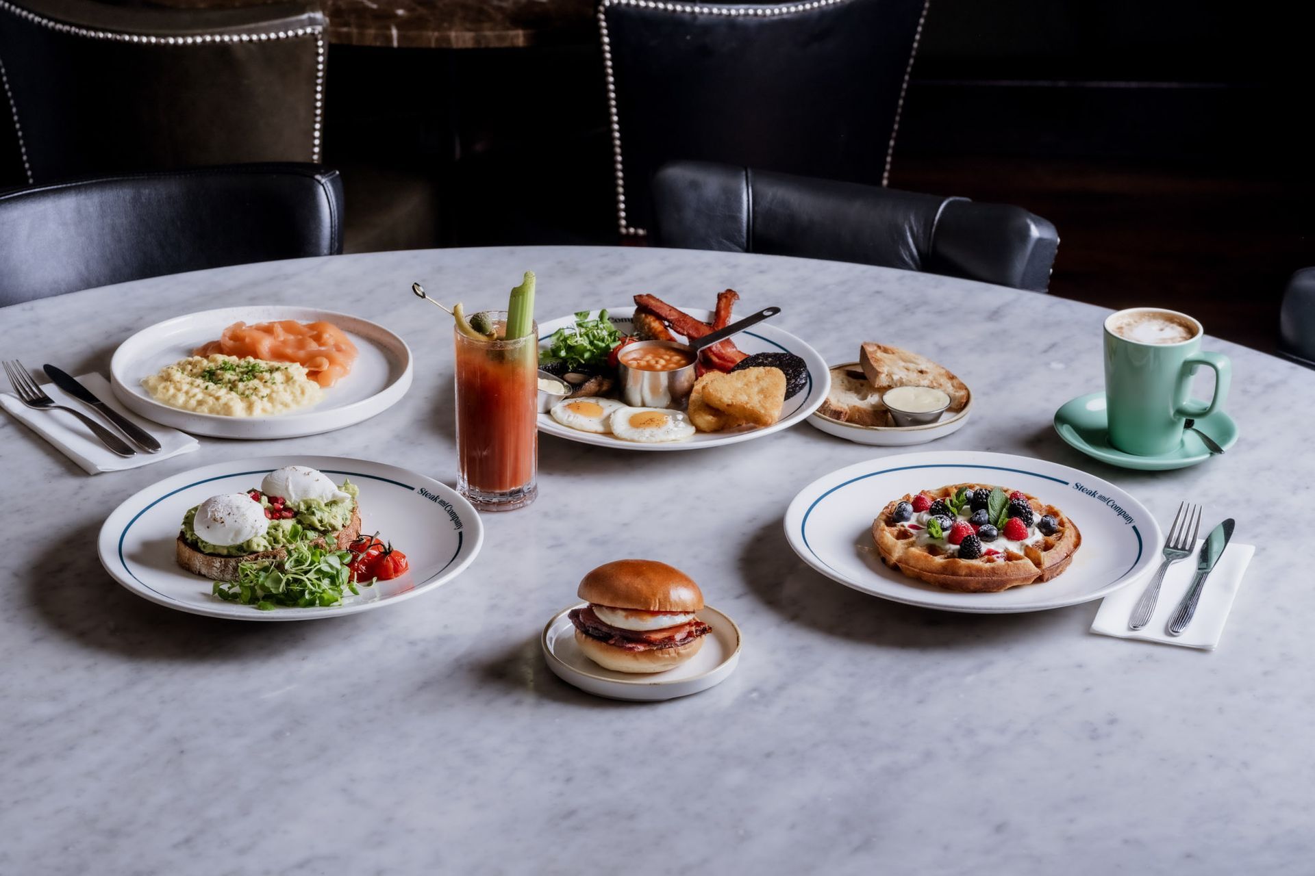 Breakfast spread on a round table: plates of food, cocktail, coffee. Dark setting.