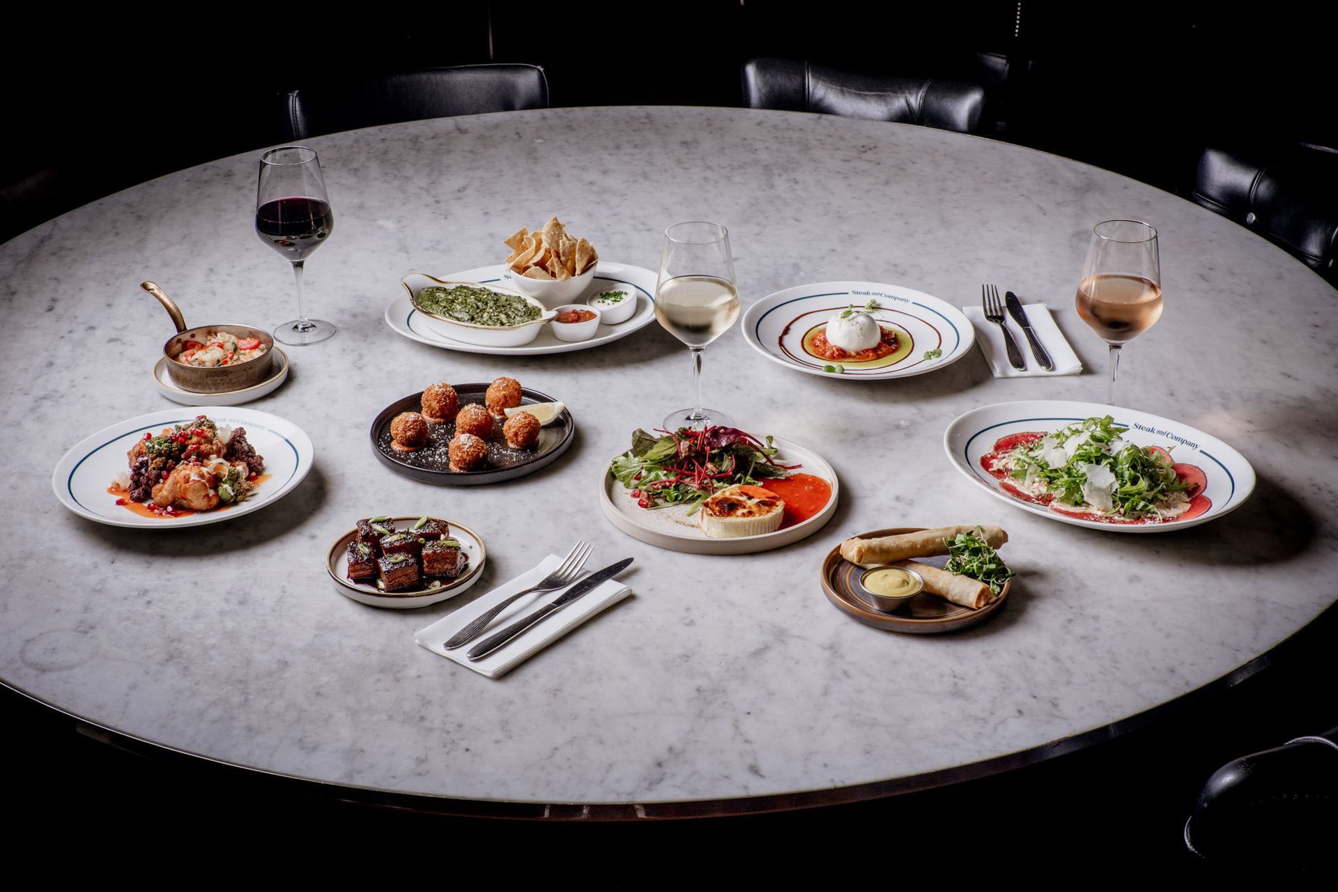 A large round table with an assortment of food and drinks: several plates of appetizers, wine glasses.