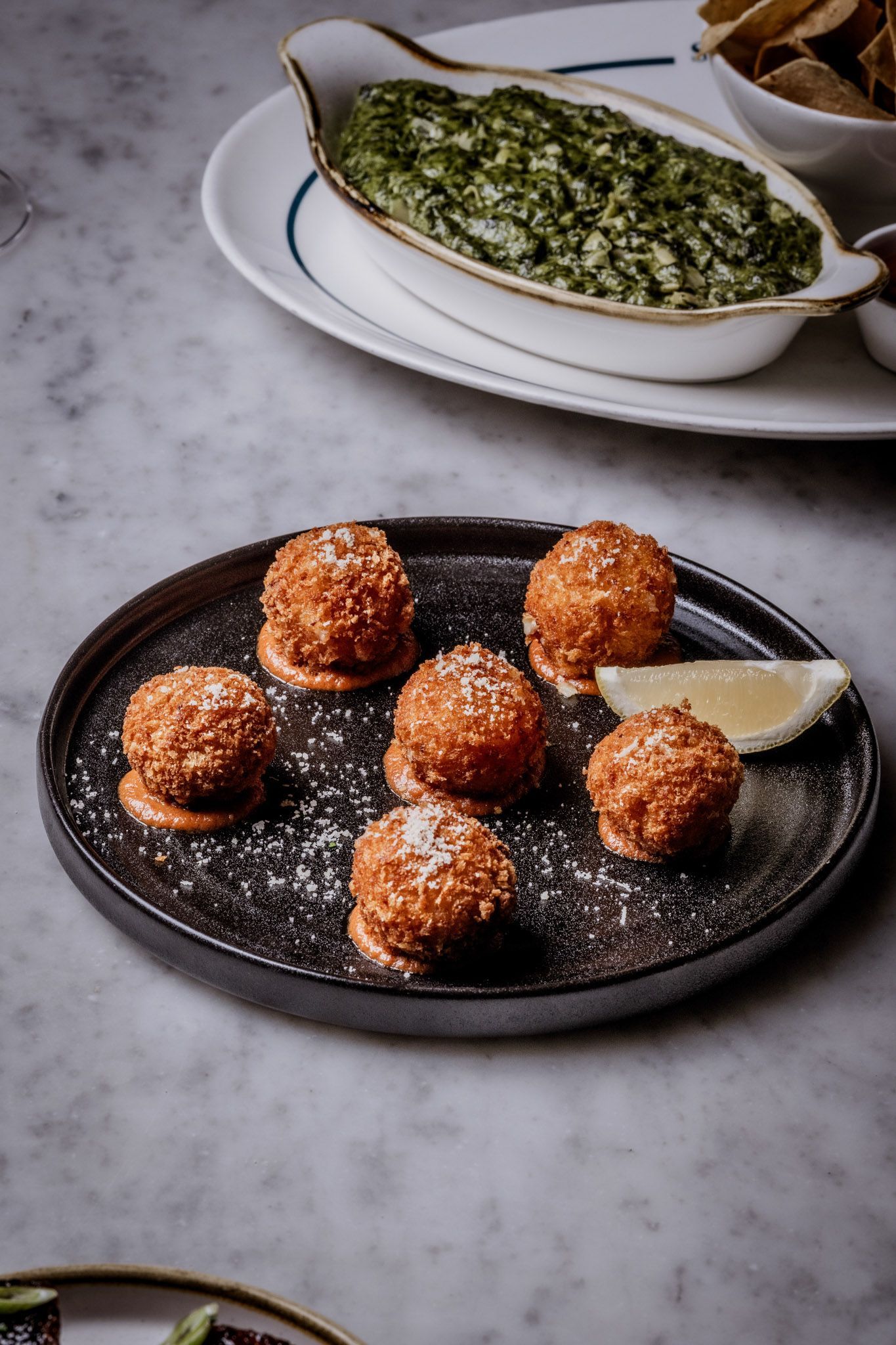 Fried food balls on a black plate with spinach dip and lemon slice on a white marble surface.