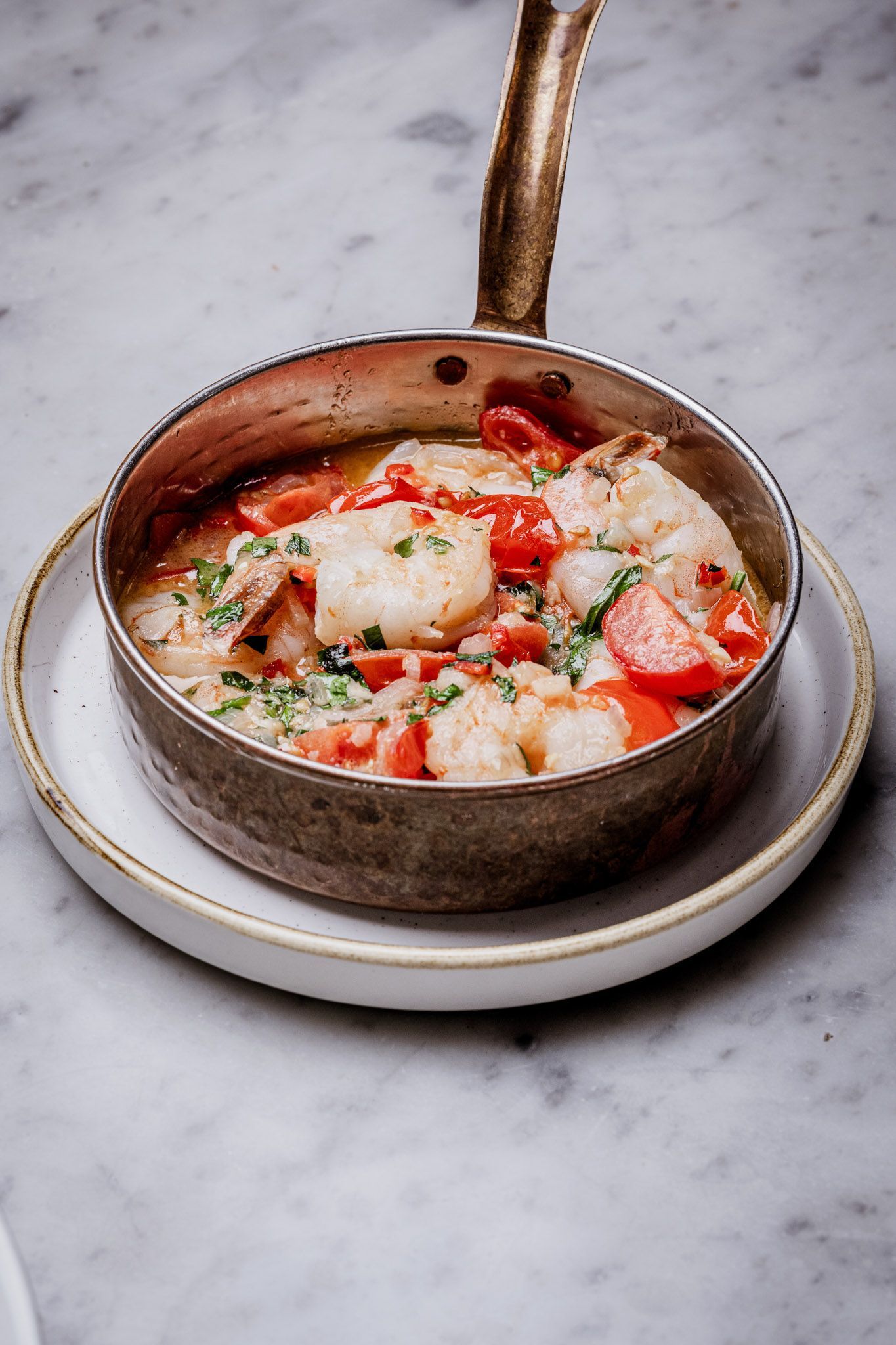 Shrimp and tomato dish in a small silver pan on a white plate, marble surface.