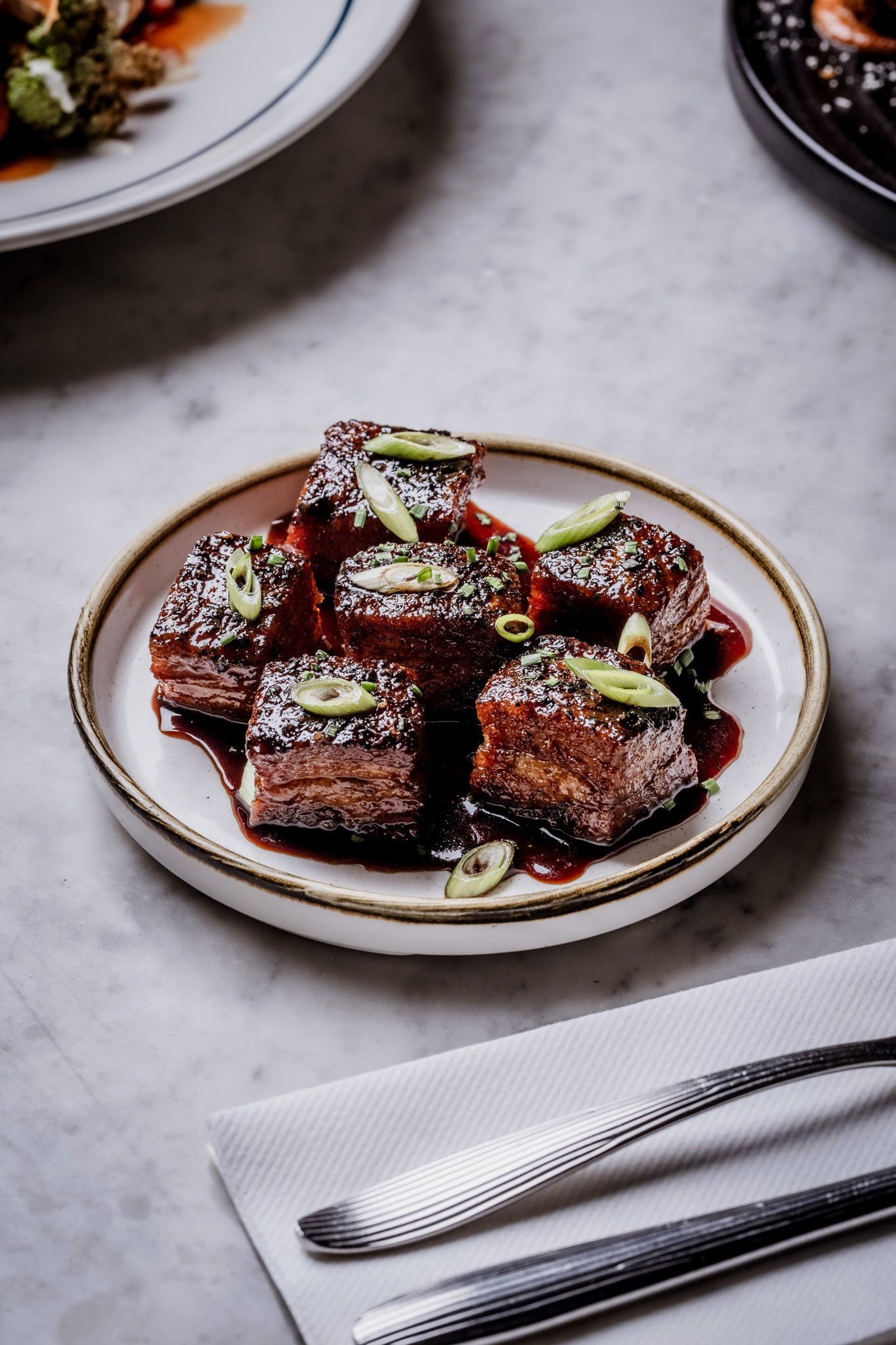 Plate of glazed pork belly cubes, garnished with green onions, on a marble table with cutlery.