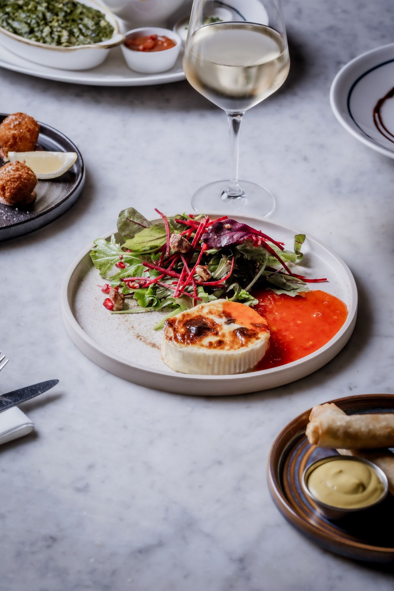 Plate with salad, baked item, and wine glass on a marble table with other dishes and condiments.