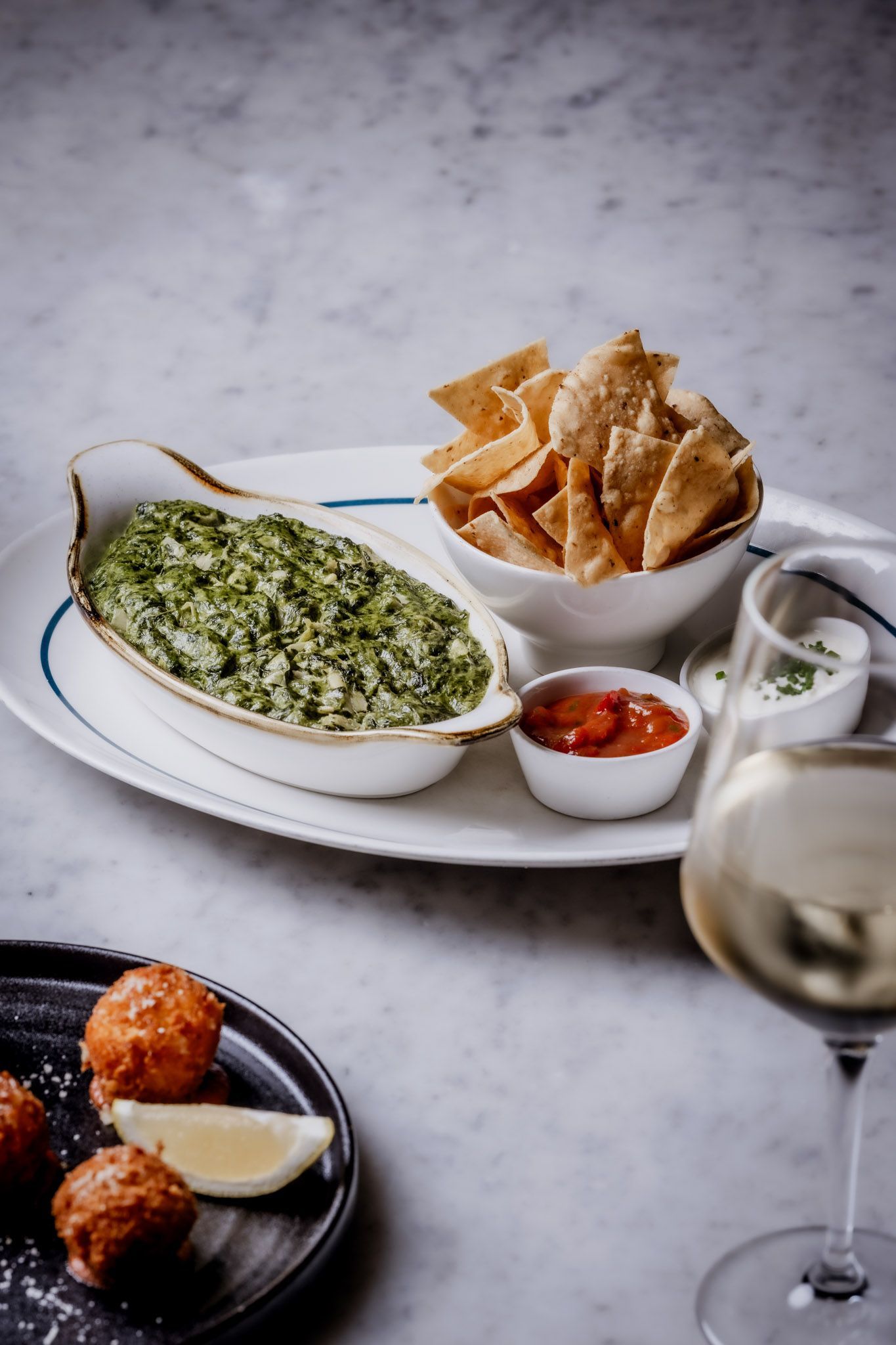 Spinach dip with chips, salsa, and wine on a white platter; fried balls and lemon slice on black plate.