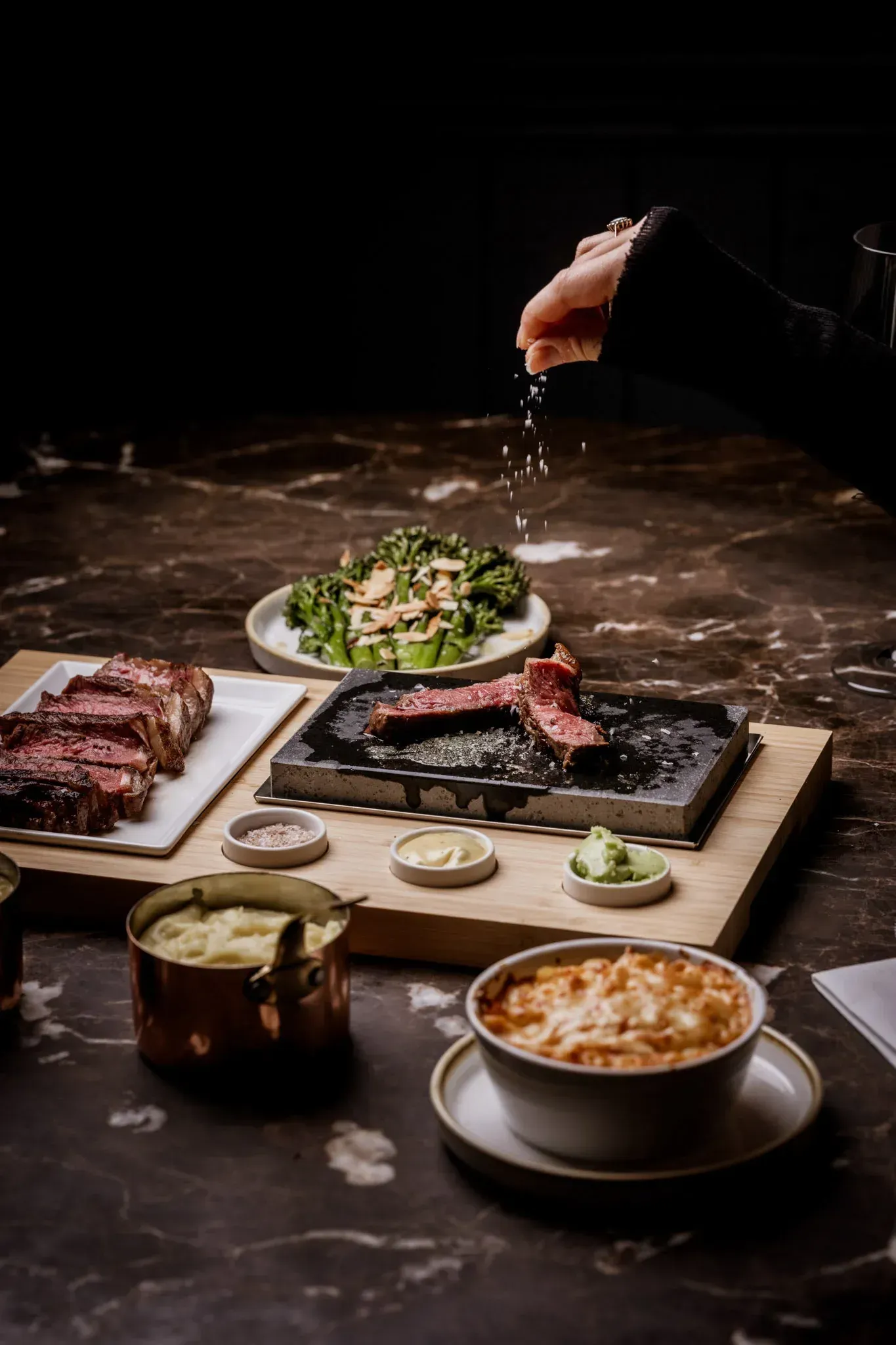 A person salting steak on a wooden board. Side dishes and other steaks are on a marble table.