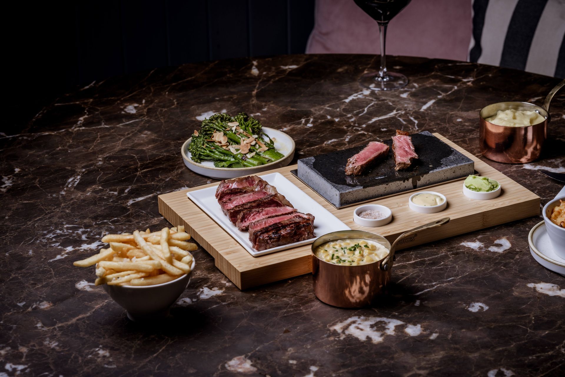 Close-up of a person slicing a cooked steak on a stone slab on a wooden tray with side dishes.