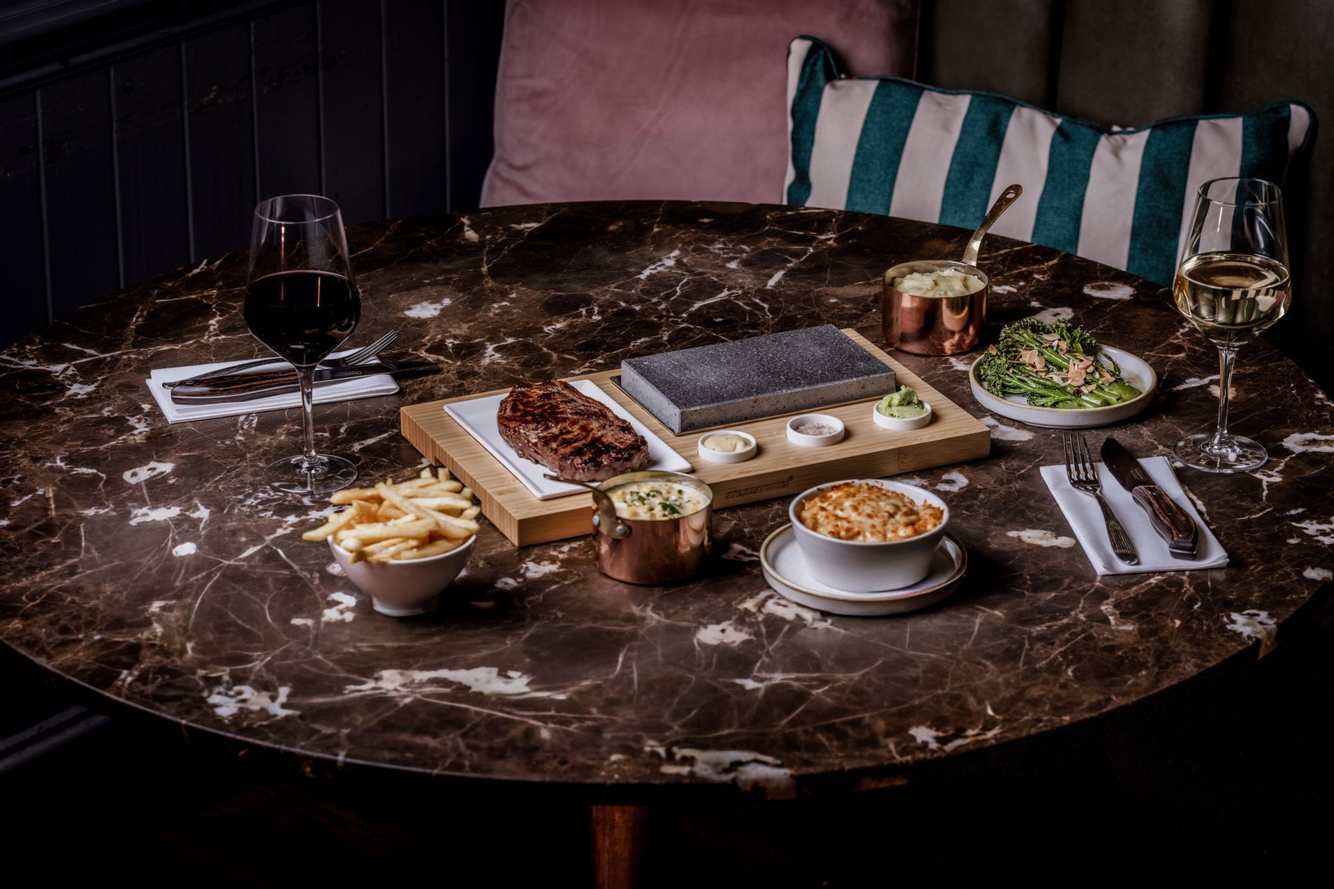 Round marble table set for a meal: steak on a hot stone, sides, wine glasses, and a salad.