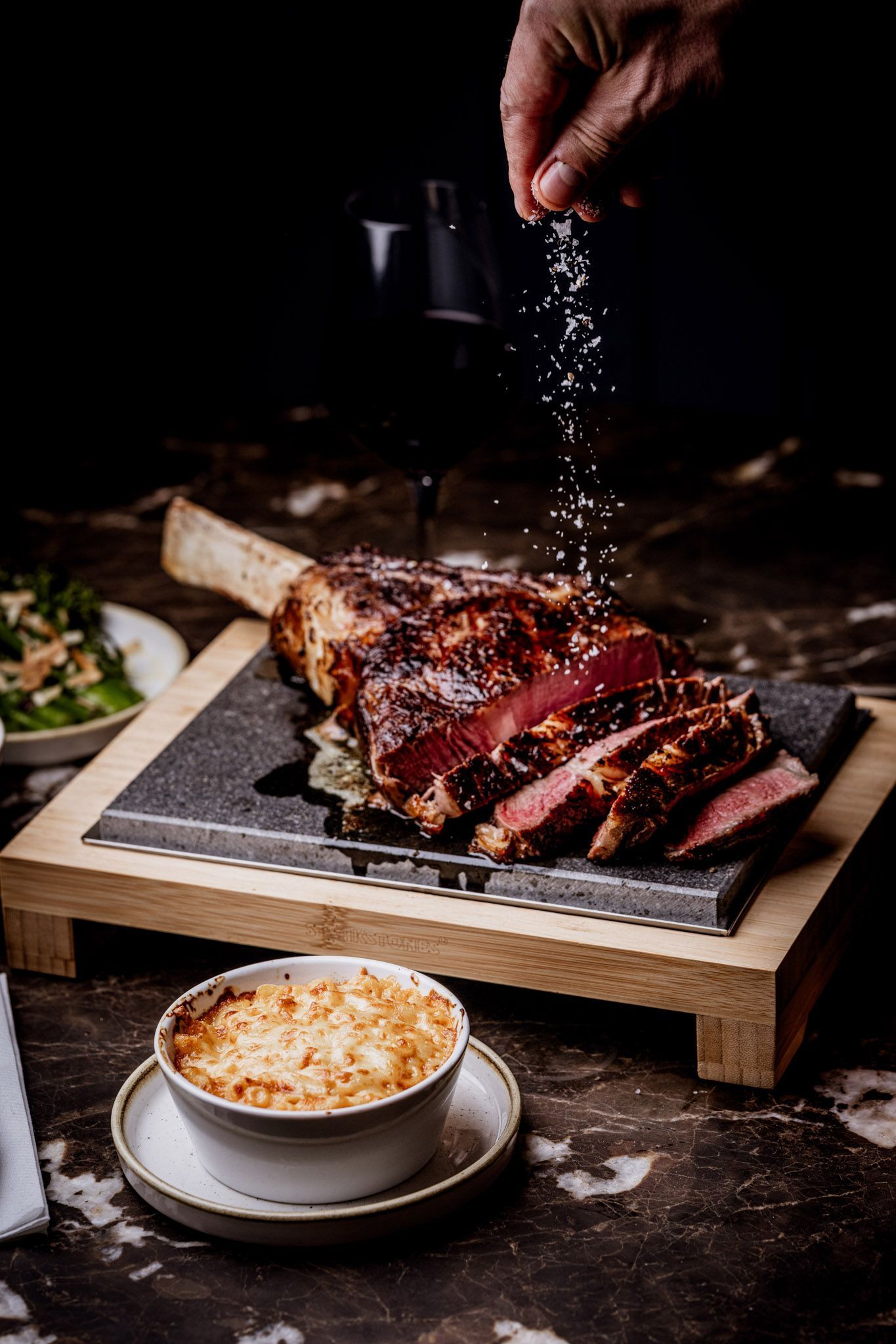 A hand sprinkling salt onto a sliced, cooked tomahawk steak on a wooden board. Side dishes complete the meal.