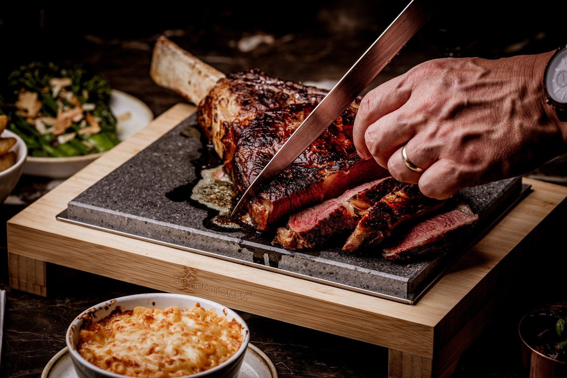 Man slicing a large steak on a wooden board; side dishes in view.