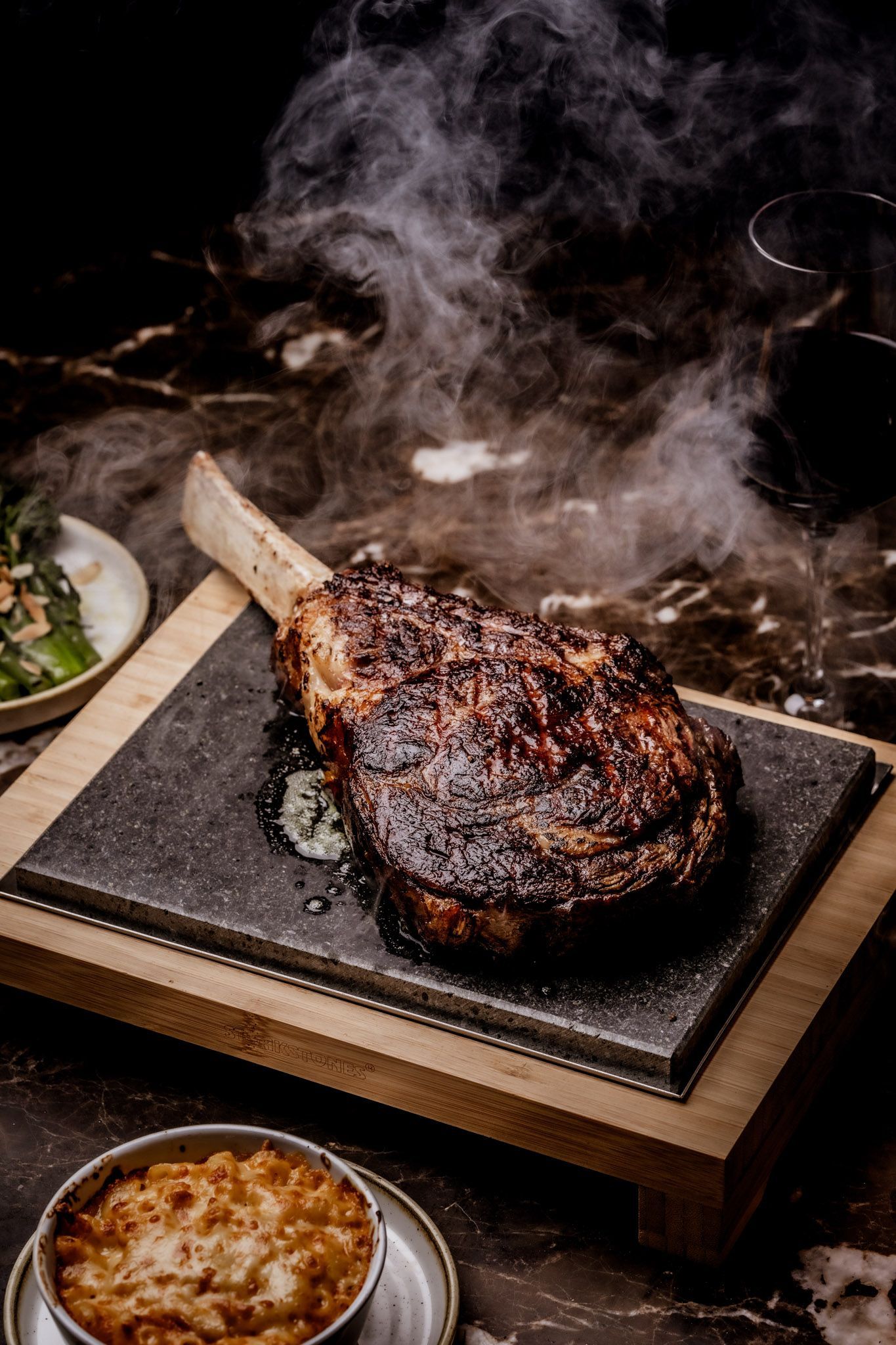 Smoked tomahawk steak on a hot stone with sides of greens and mac and cheese.