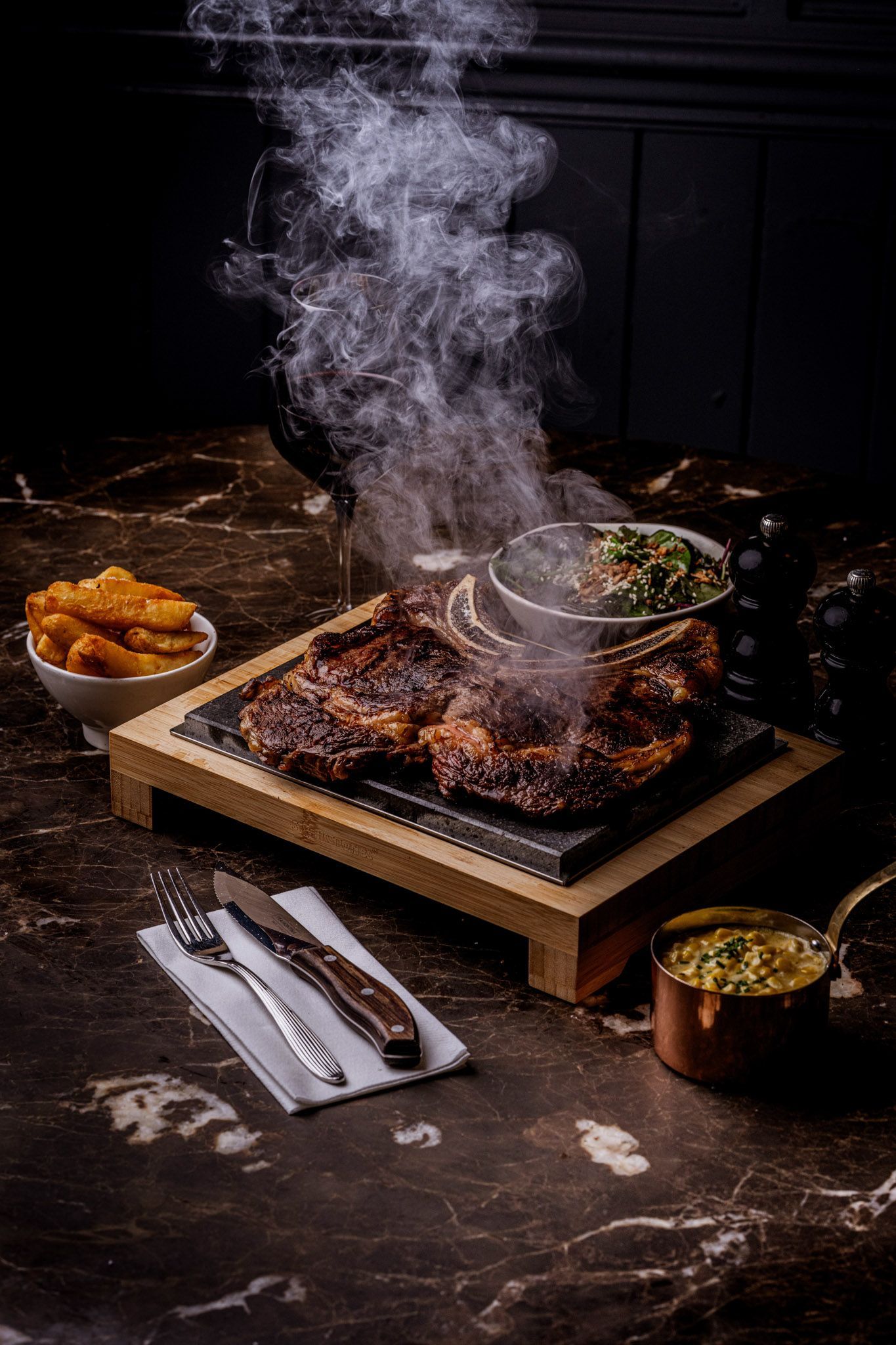 Steaming steak served on a hot stone with sides of fries and a green vegetable dish.