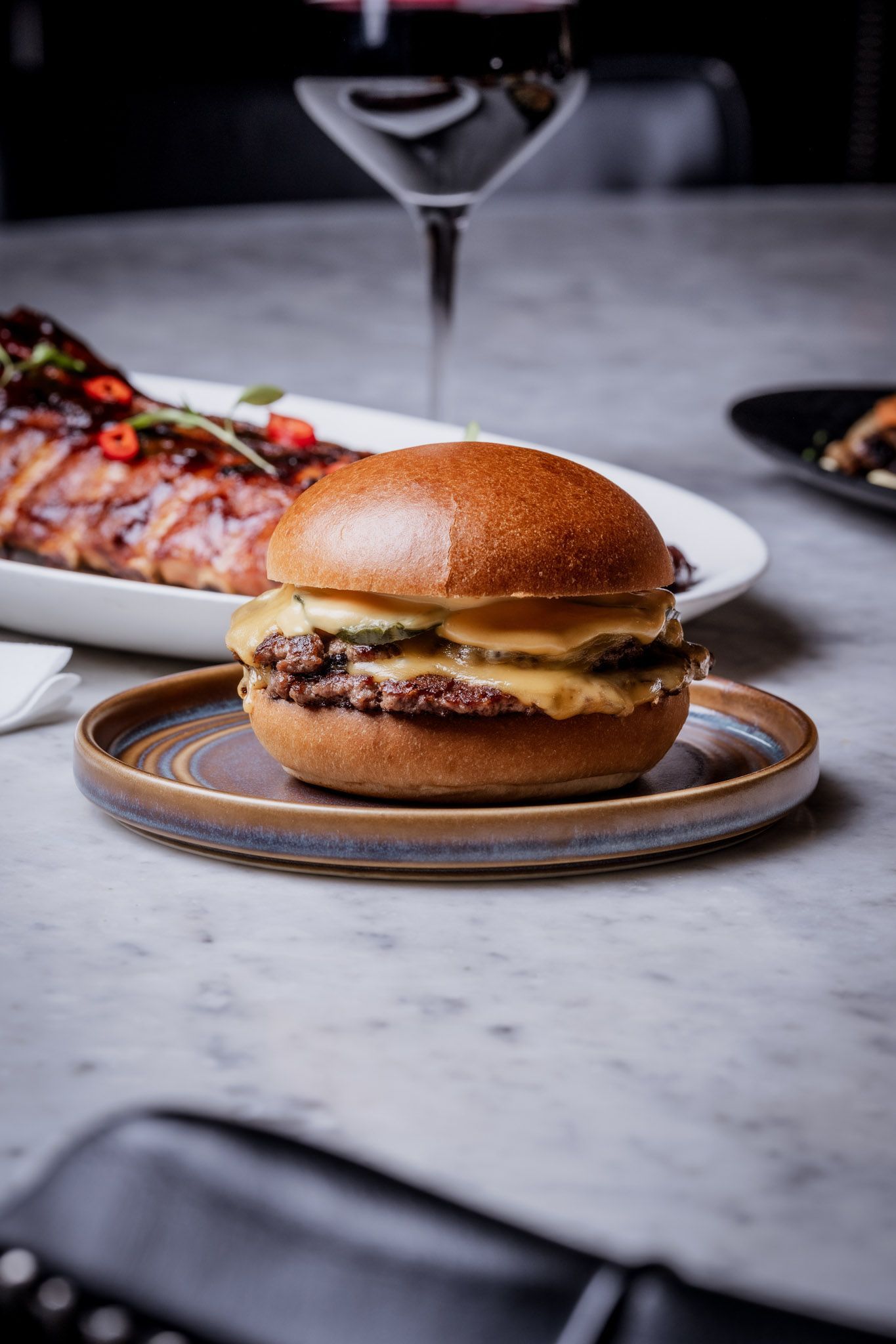Cheeseburger on a plate with blurred background of food, wine glass, and table.