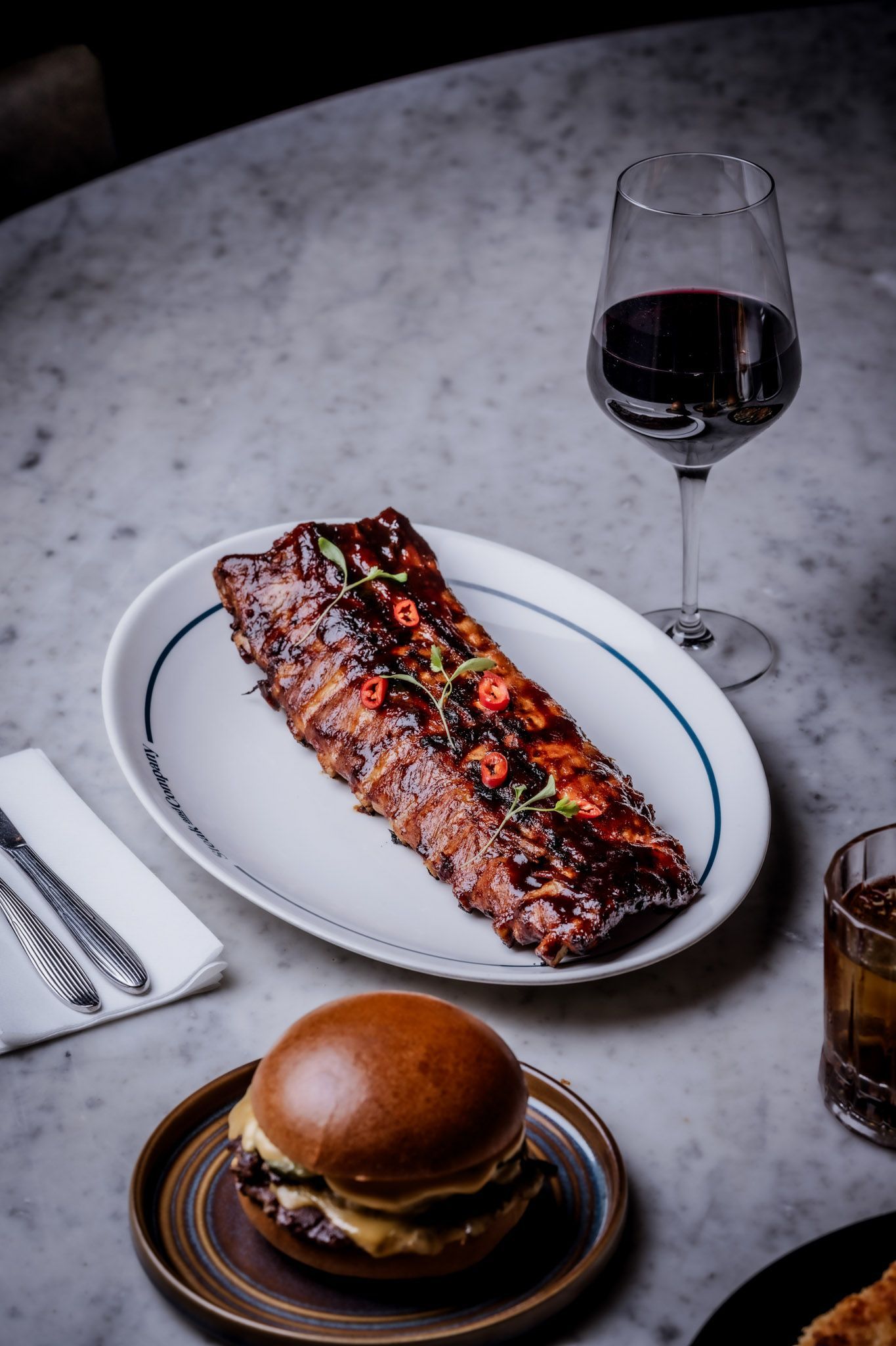Ribs on a plate with a burger, a glass of red wine, and a cocktail on a marble table.