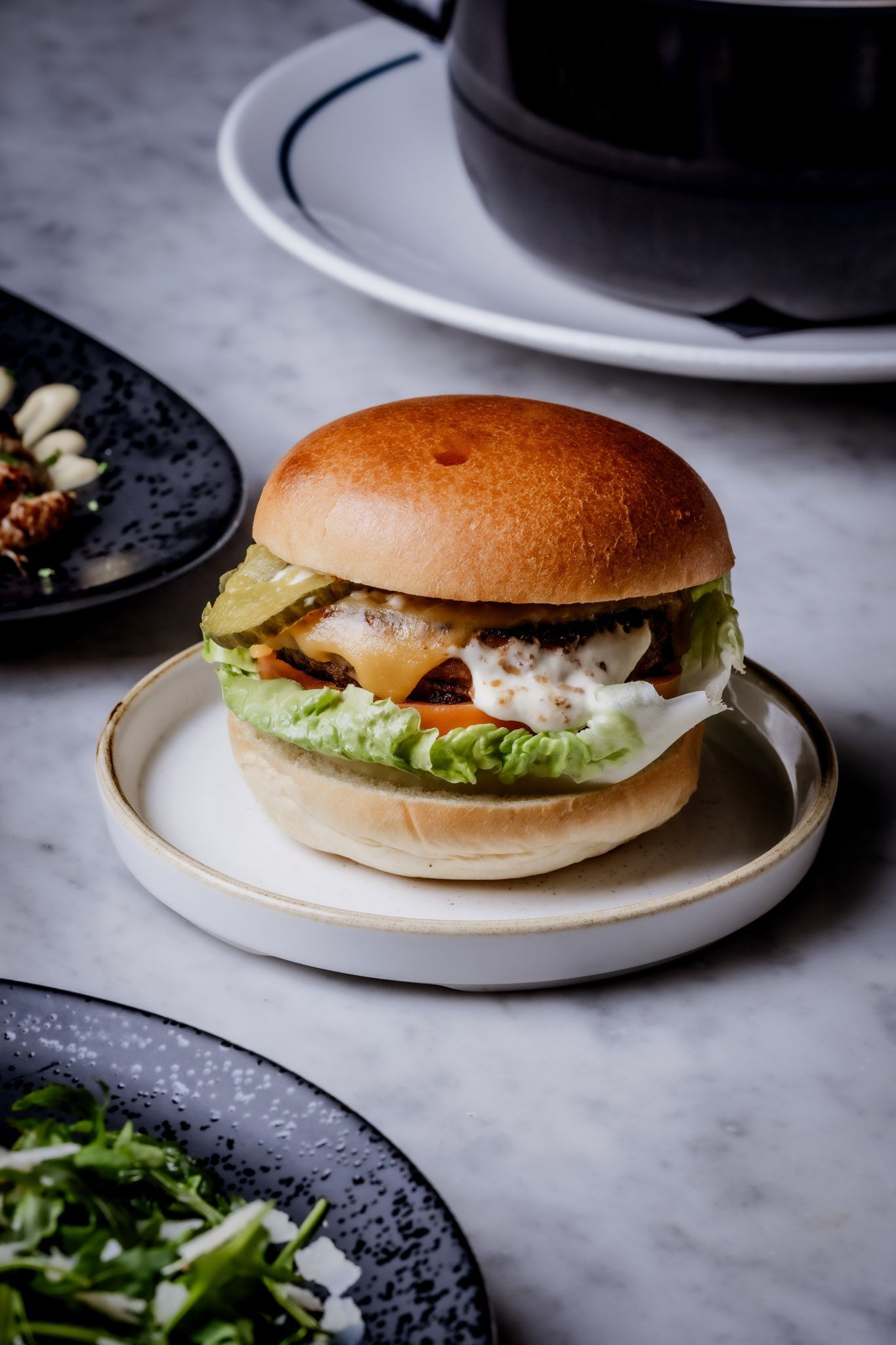 Burger on a small white plate, lettuce, pickles, and sauce visible. Other dishes and a cup are nearby.