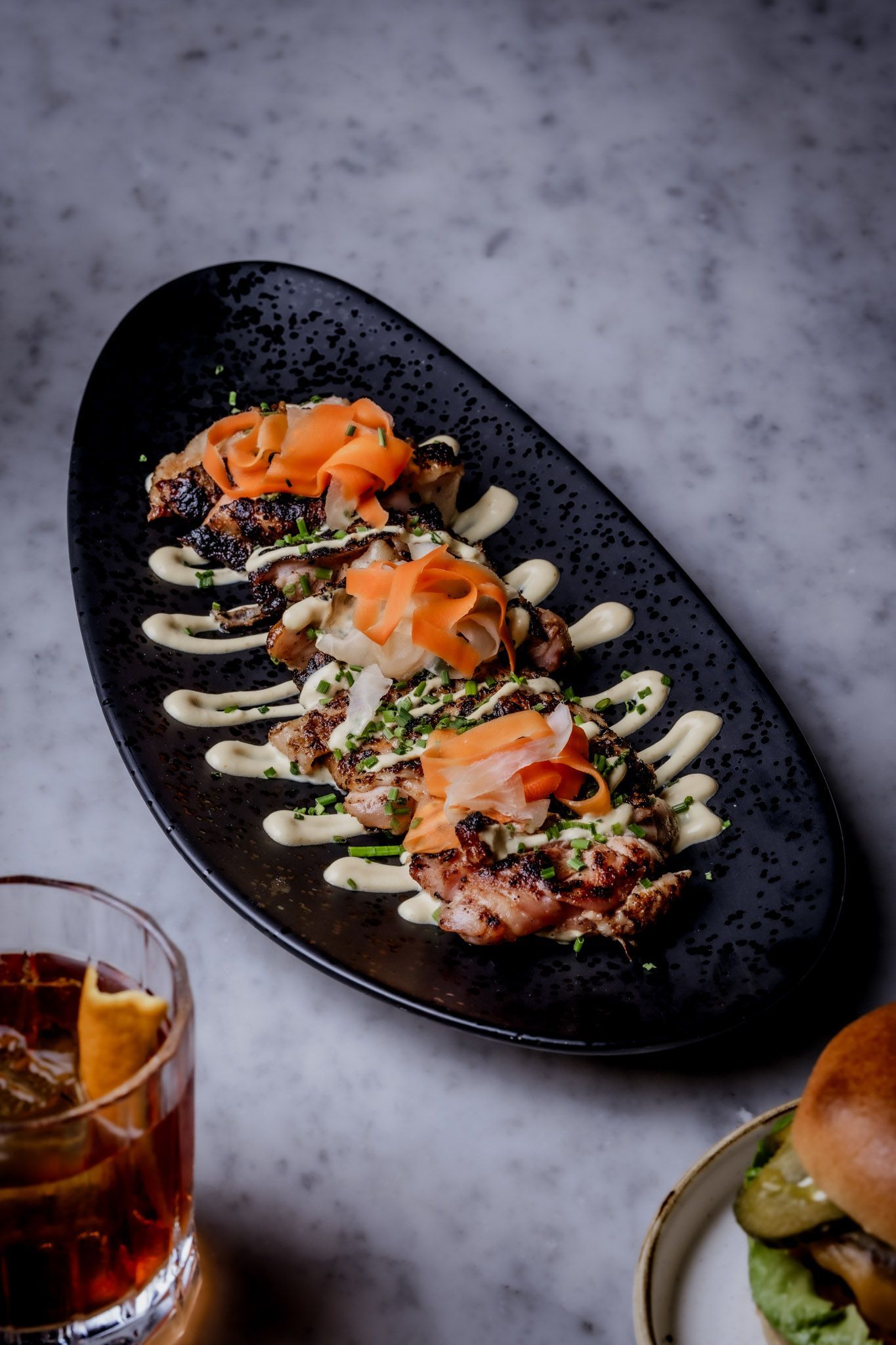Appetizer on a black oval plate with salmon, sauce drizzle, and a cocktail in the foreground.