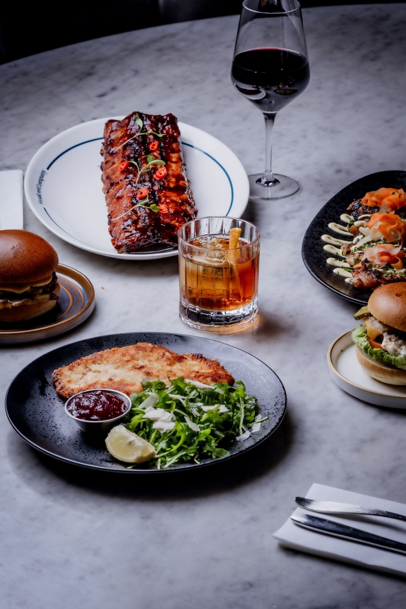 Assortment of dishes on a marble table: ribs, burger, schnitzel, sushi-style dish, cocktail, wine glass.
