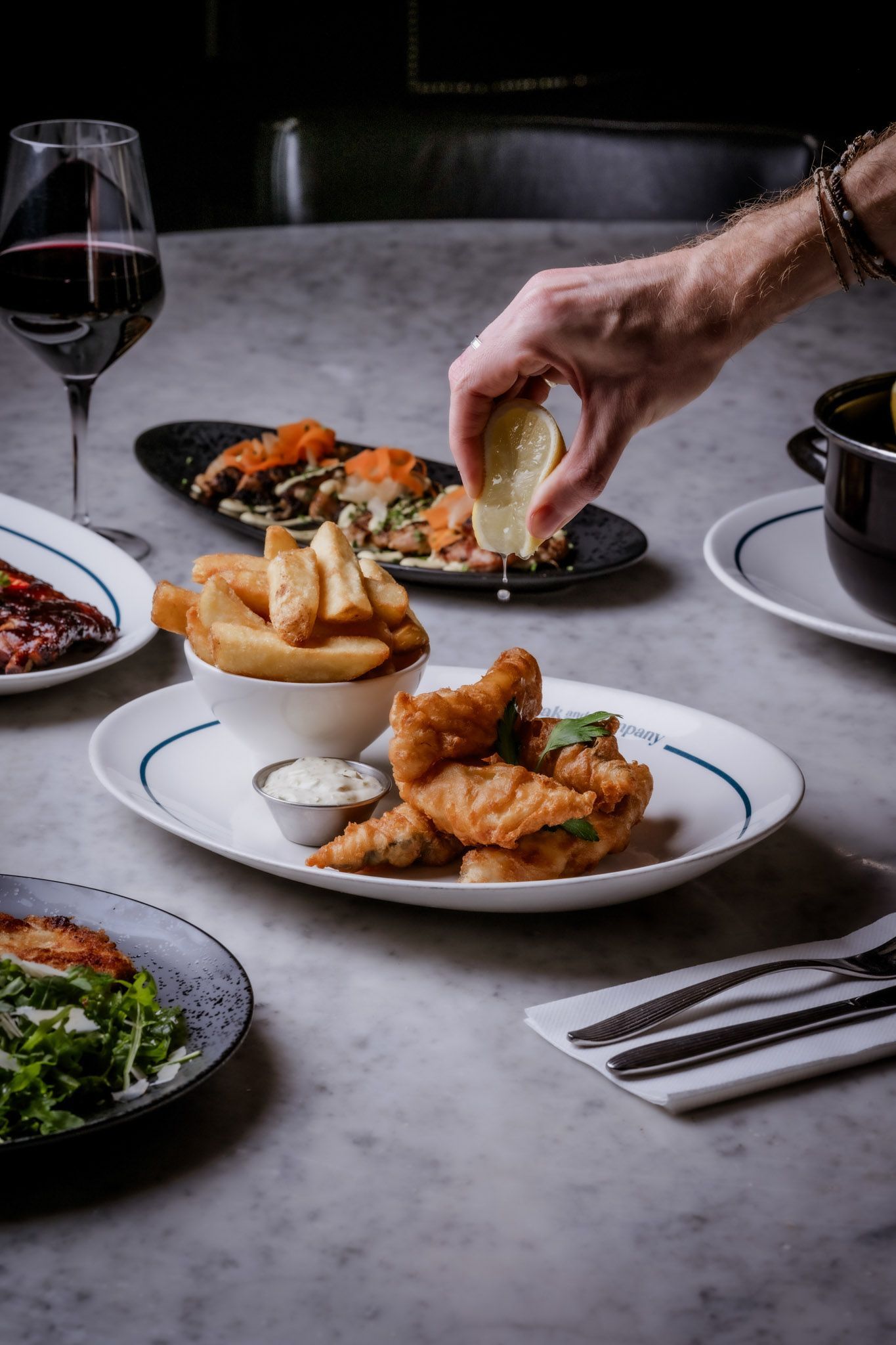 Person squeezing lemon over fried fish and chips, other dishes, and wine on marble table.