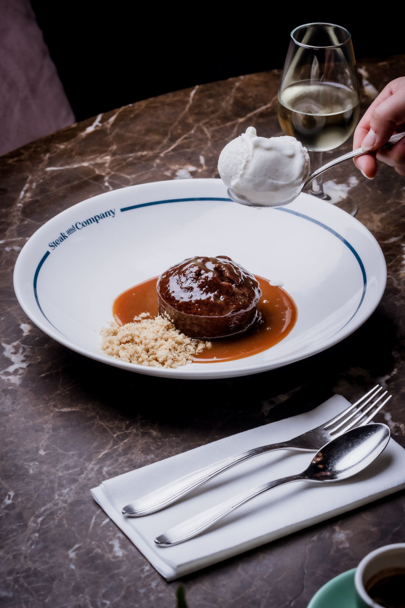 Sticky toffee pudding with ice cream, sauce, and crumble on a plate, being served next to flatware and wine.