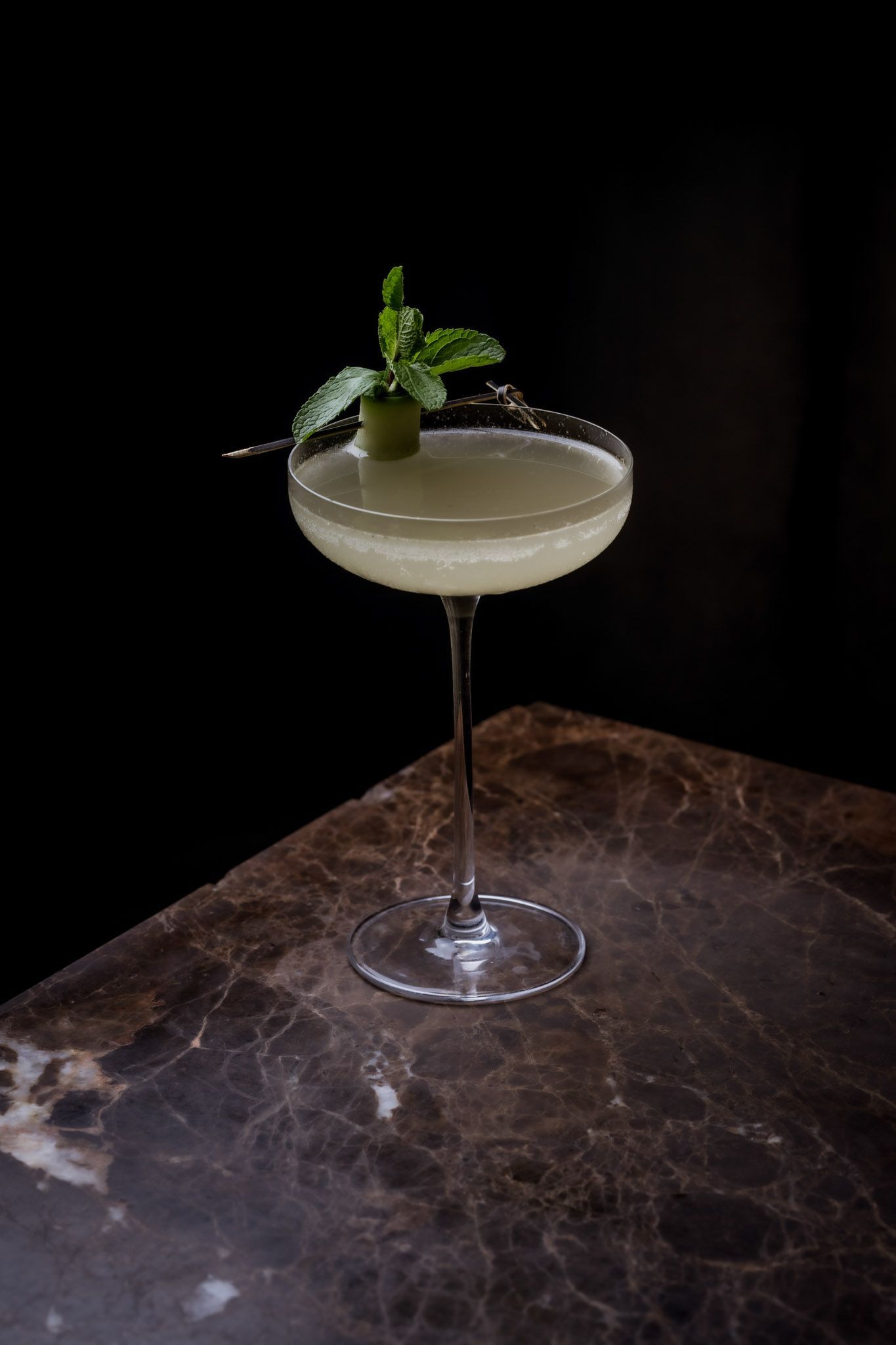 Elegant cocktail in a stemmed glass, garnished with mint, on a marble surface, black background.