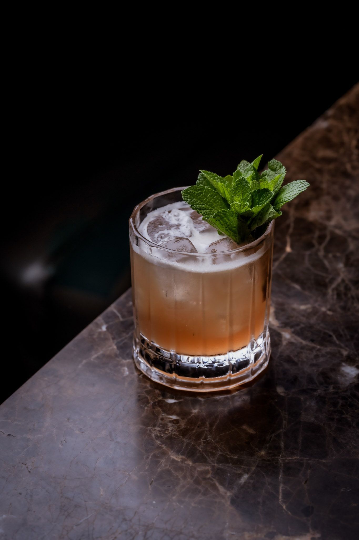 Cocktail in a glass with mint garnish on a marble surface, against a dark background.