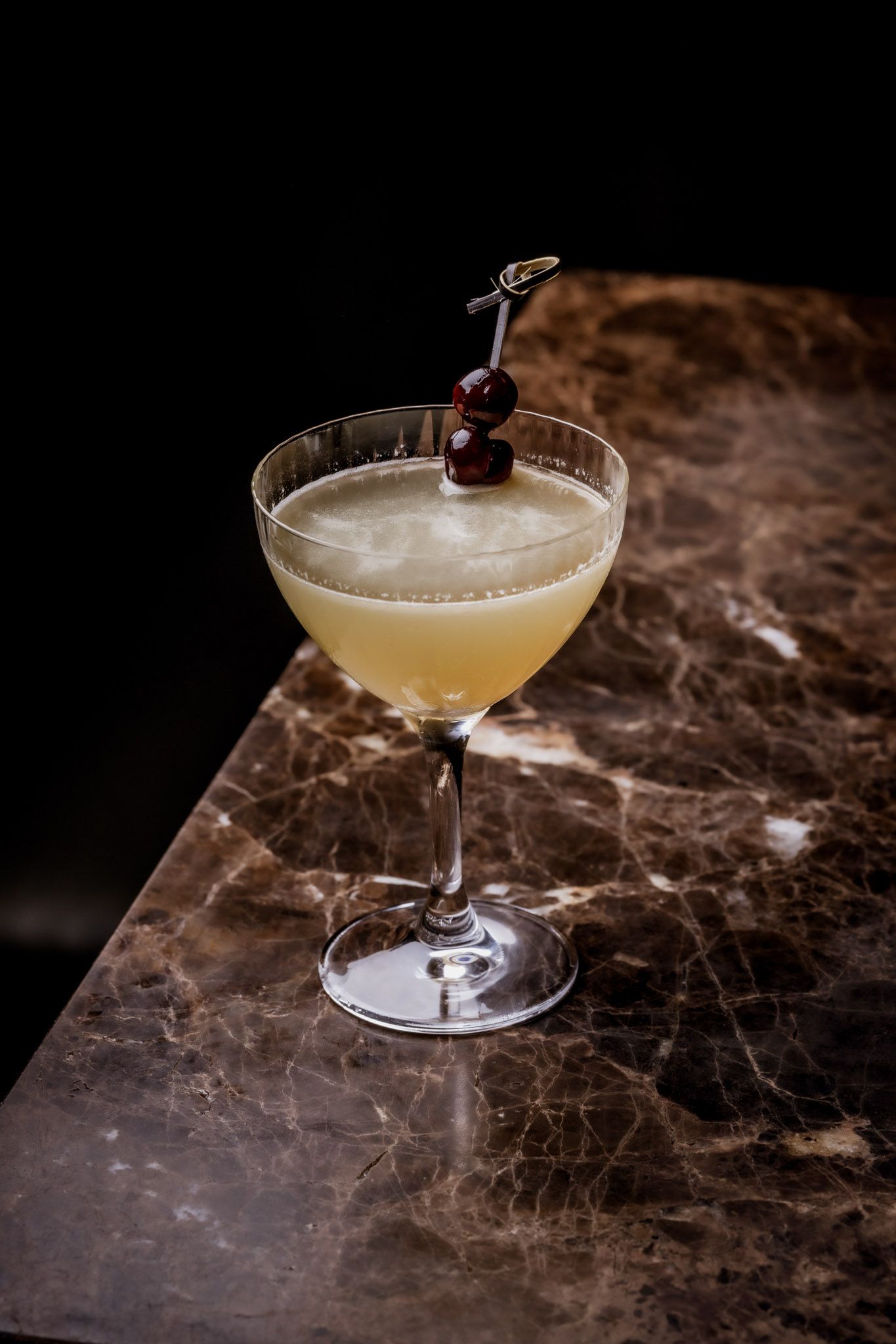 Cocktail in a stemmed glass, garnished with cherries, on a marble-patterned surface.