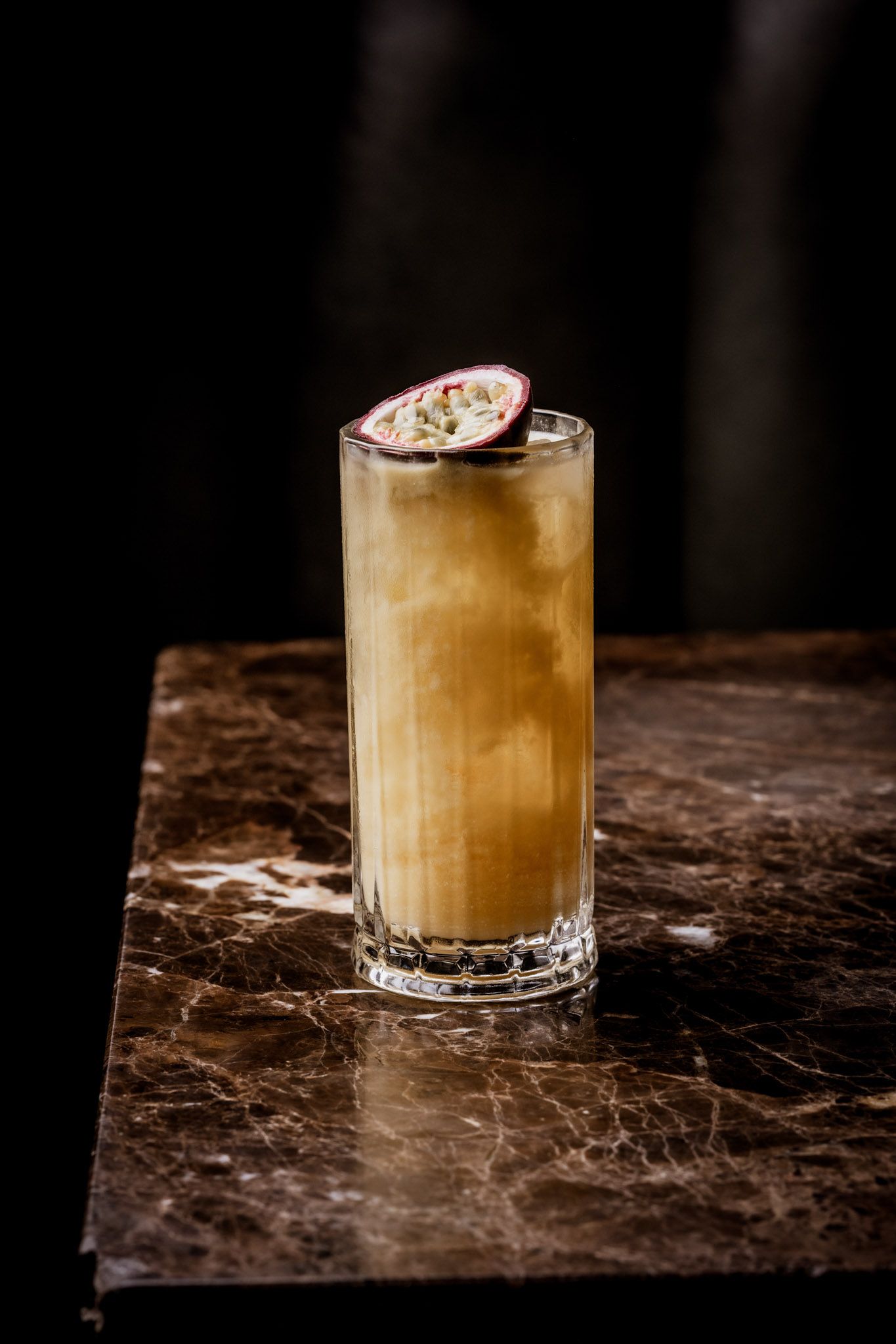 Cocktail in tall glass, garnished with passion fruit, on a marbled surface. Dark background.