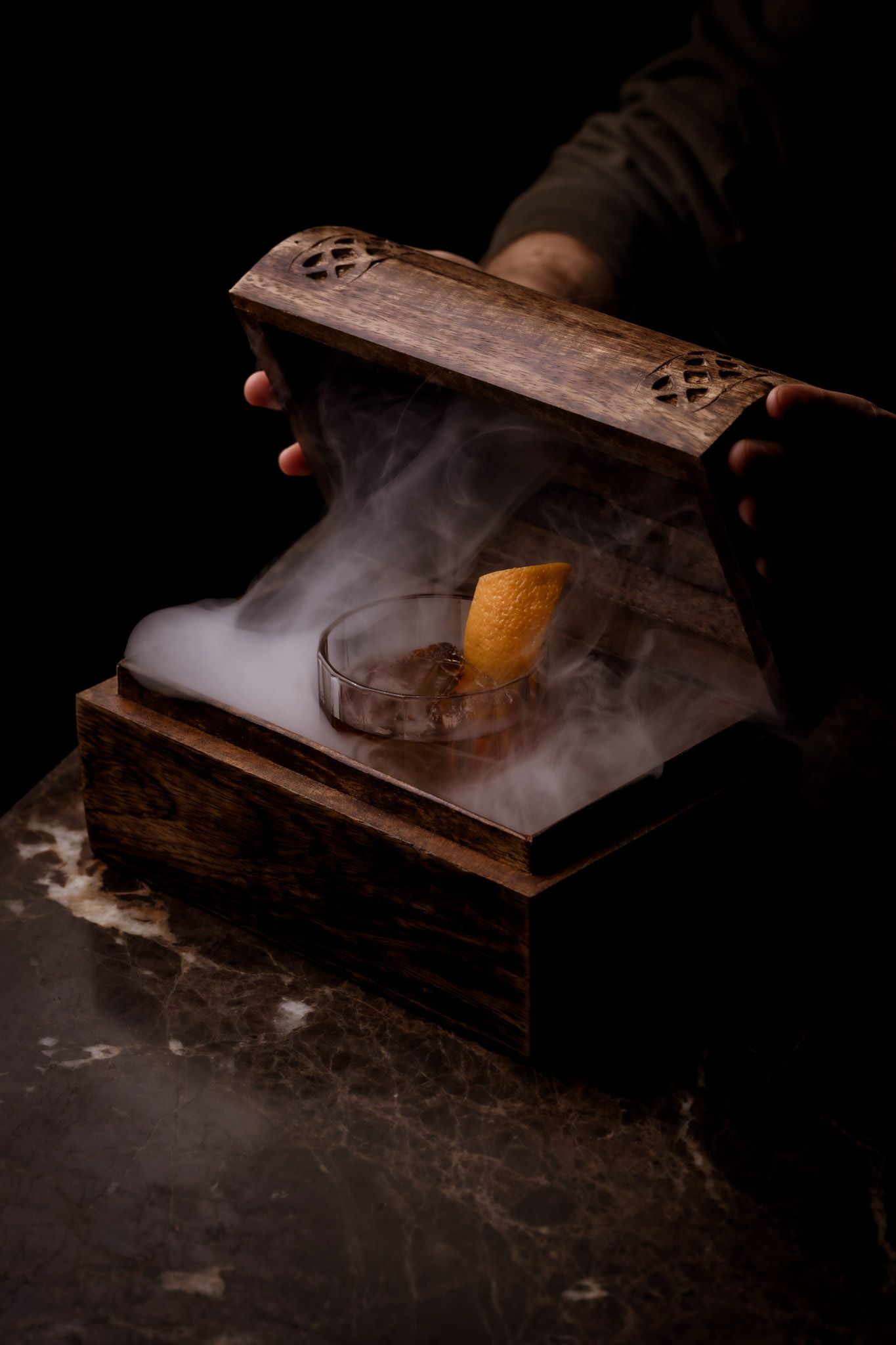 Cocktail in a wooden box with smoke, being opened by hands.