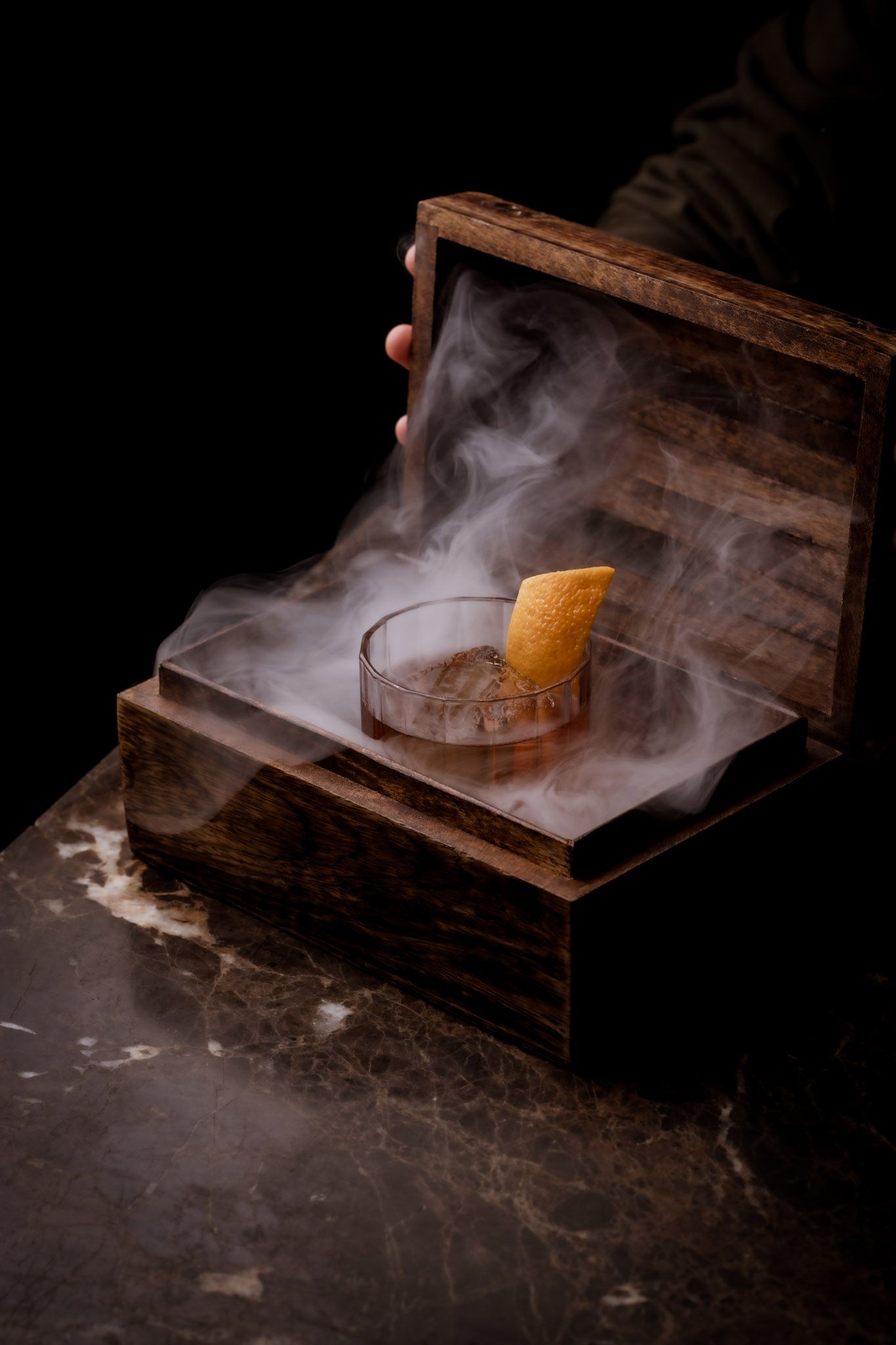 Cocktail in a wooden box with smoke, topped with an orange peel, on a marble table.