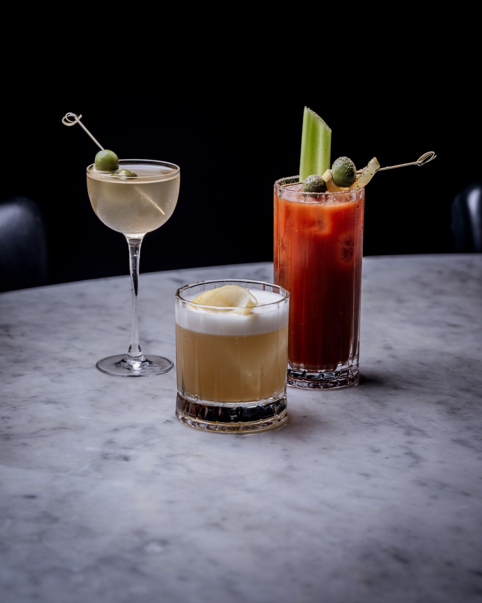 Three cocktails on a marble table: a martini, Bloody Mary, and sour.